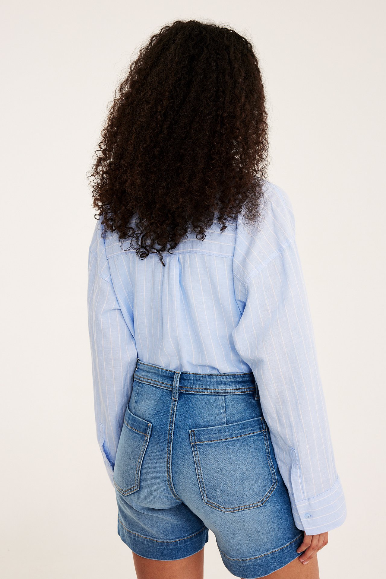 Nainen selin: vaaleansiniset denimsortsit taskuilla ja raidallinen vaaleansininen paita.