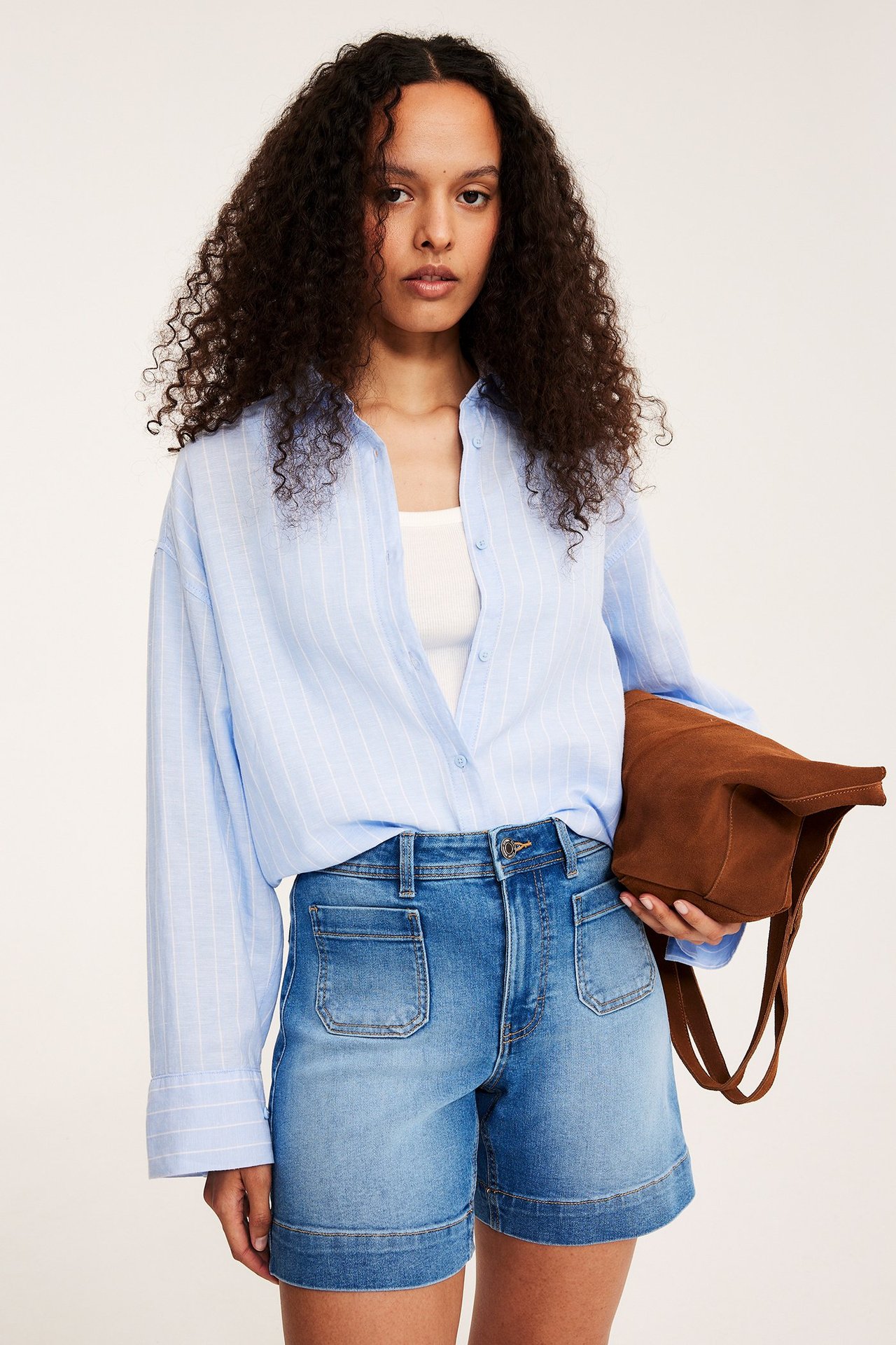 Kvinna bär ljusblå jeansshorts med utanpåliggande fickor, vit topp, randig blå skjorta, håller brun väska.