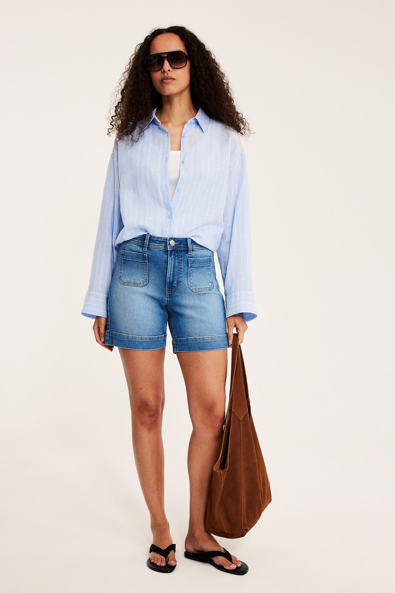 Frontvy av kvinna i ljusa denimshorts med fickor, blårandig skjorta, solglasögon, håller brun mockaväska.