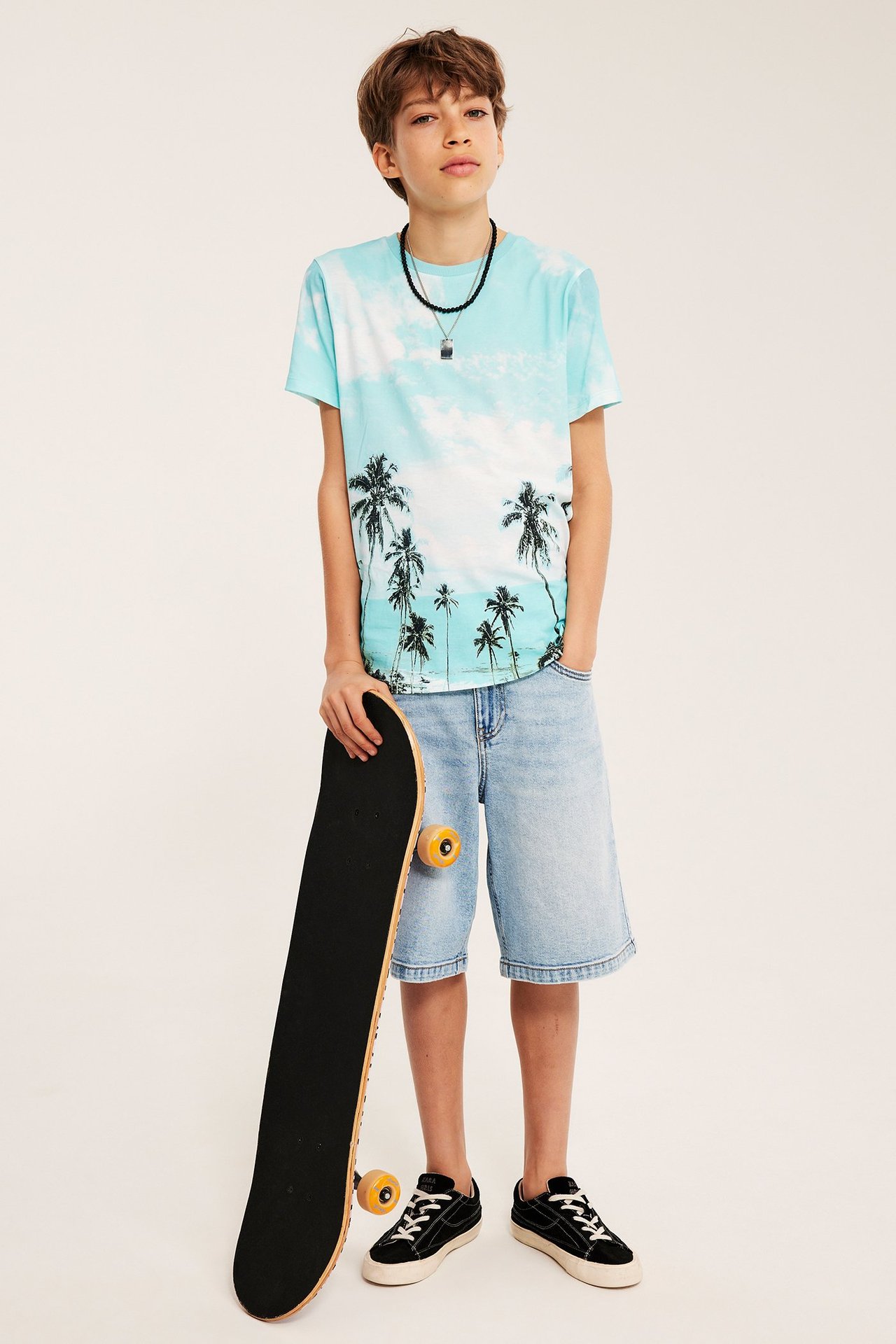 Framifrån: Barn i offwhite, ljusblå T-shirt med palmmönster, jeansshorts, svarta sneakers, håller skateboard.
