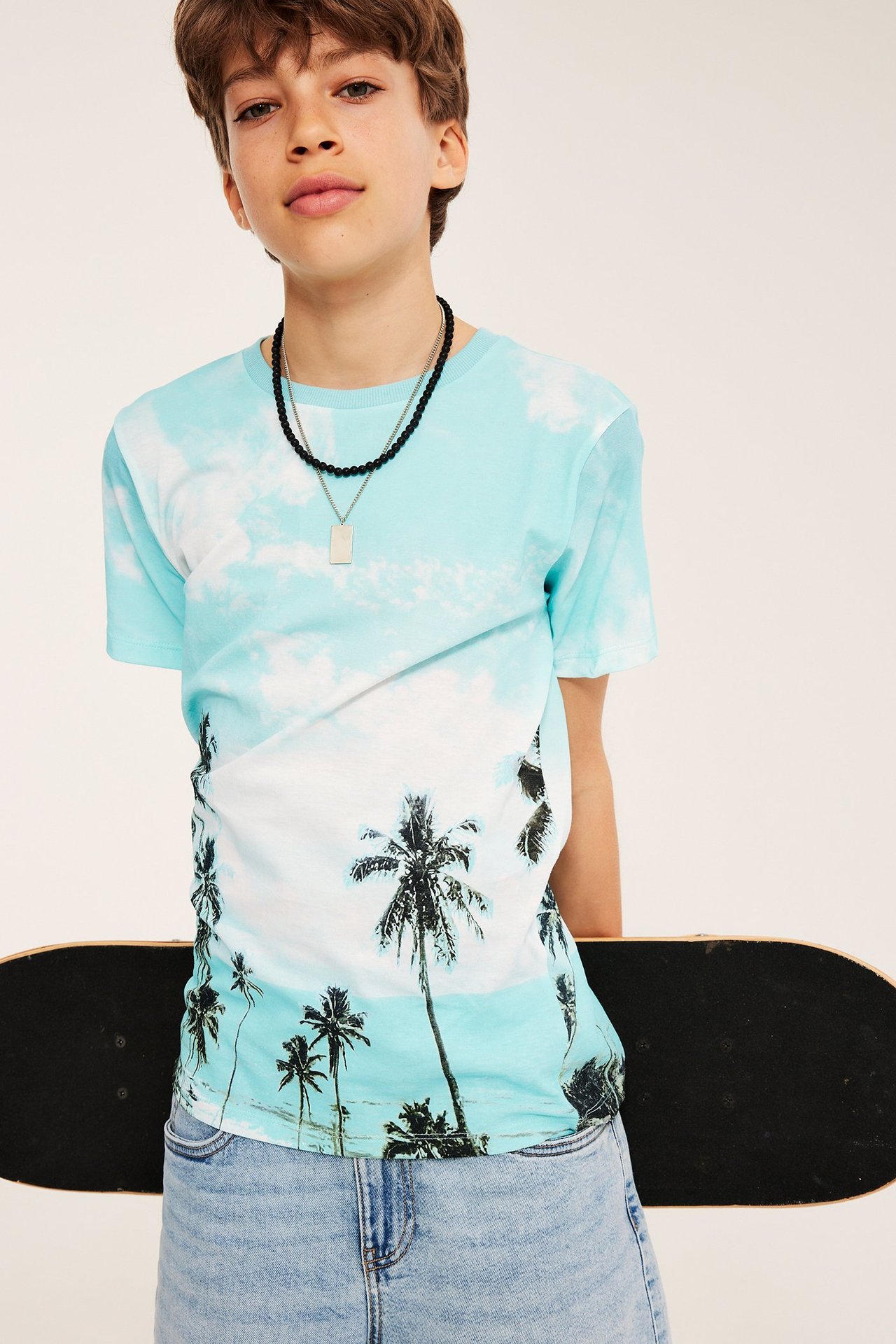 Koszulka dziecięca jasnoniebieska z nadrukiem palm, jasne szorty jeansowe i naszyjnik. Deskorolka z tyłu.