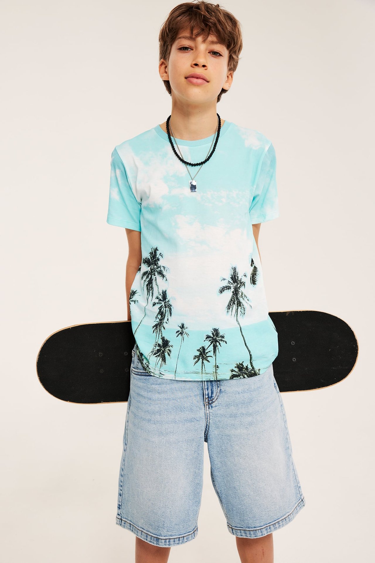 Barns frontvy: ljusblå bomull t-shirt med palmtryck, ljusa jeansshorts, svart pärlhalsband, håller skateboard.
