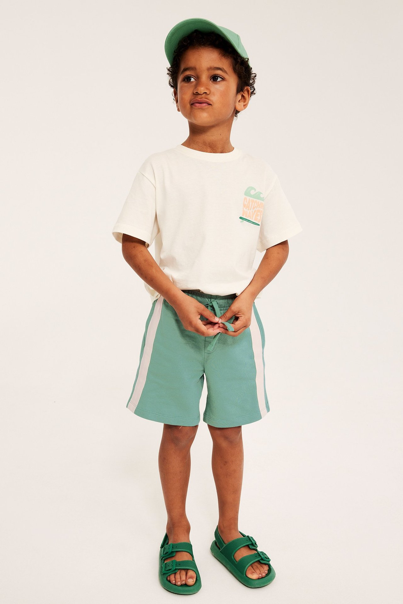 Barn i gröna jersey shorts med vita revärer, benvit t-shirt med tryck, grön keps och gröna sandaler.