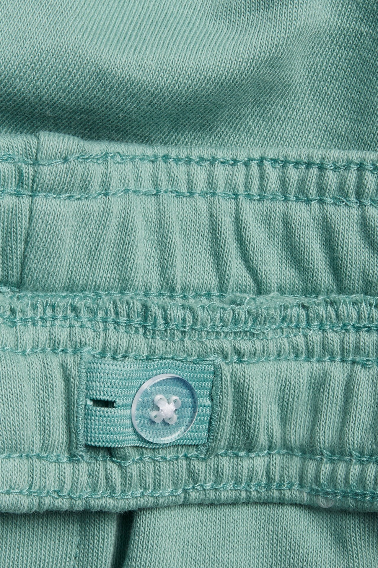 Lähikuva vihreiden collegeshortsien vyötärönauhasta säädettävällä napilla ja napinlävellä.