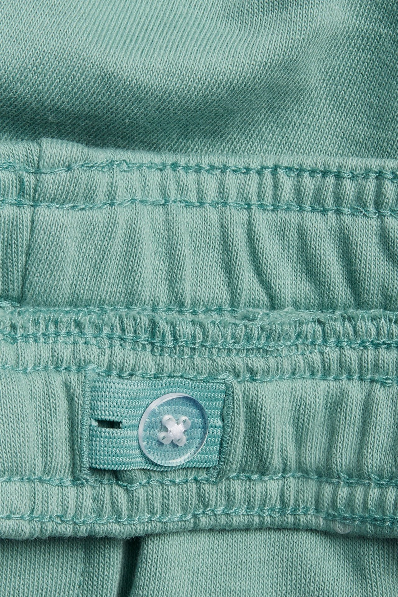 Lähikuva vihreiden collegeshortsien vyötärönauhasta säädettävällä napilla ja napinlävellä.