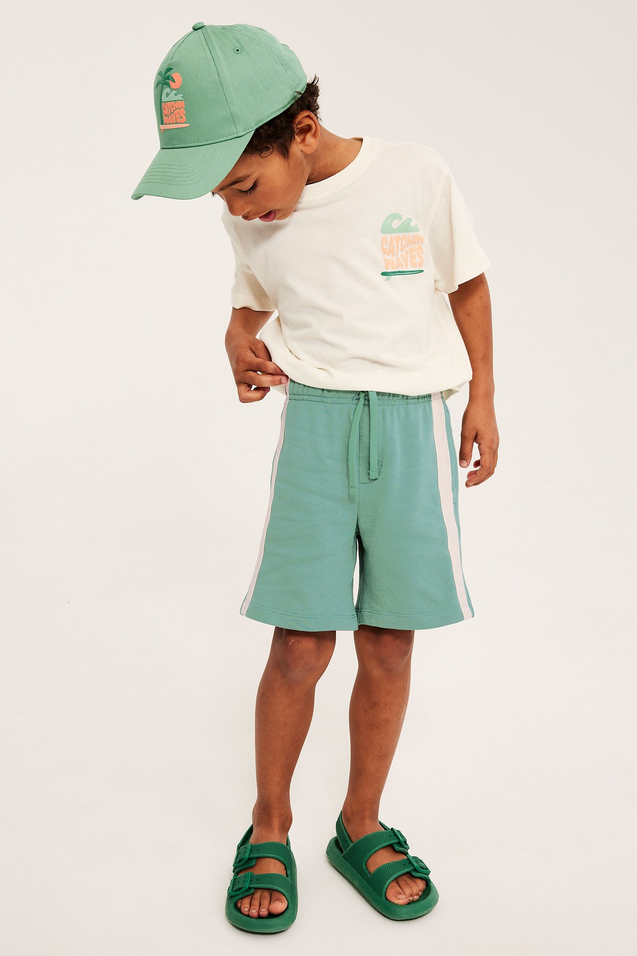Framifrån: Barn bär gröna mjukisshorts med revärer, krämfärgad grafisk t-shirt, grön keps och gröna sandaler.