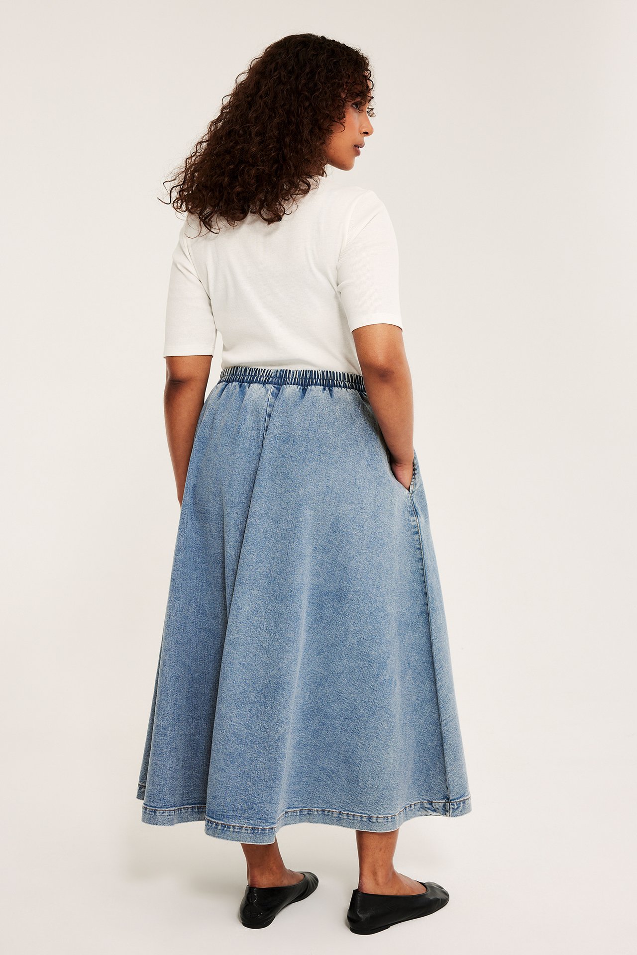 Bakifrån: kvinna i ljusblå jeanskjol med resår i midjan, sidofickor, vit t-shirt och svarta platta skor.