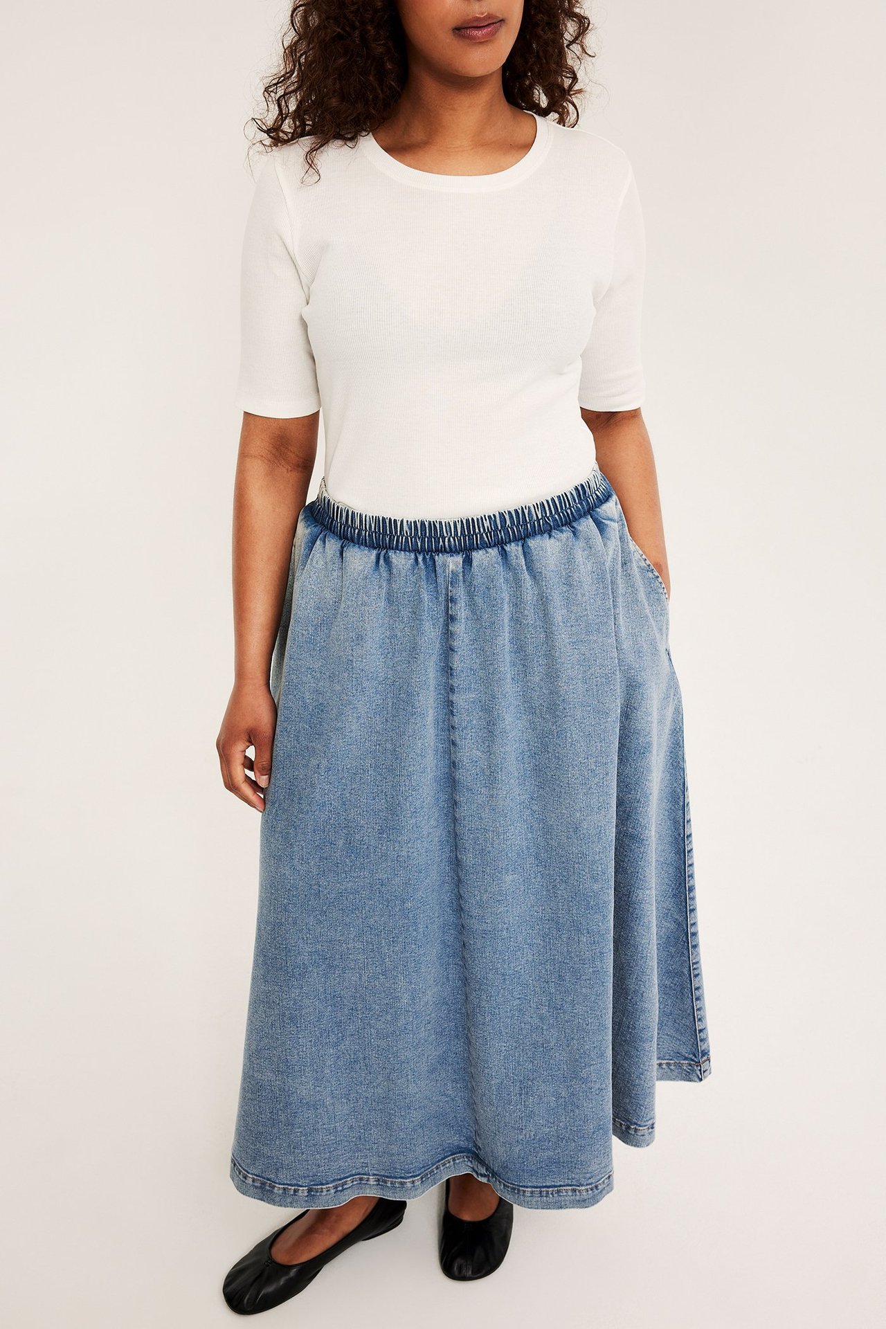 Framifrån: Kvinna i ljus jeanskjol med resår, sidofickor, vit t-shirt, svarta ballerinaskor.