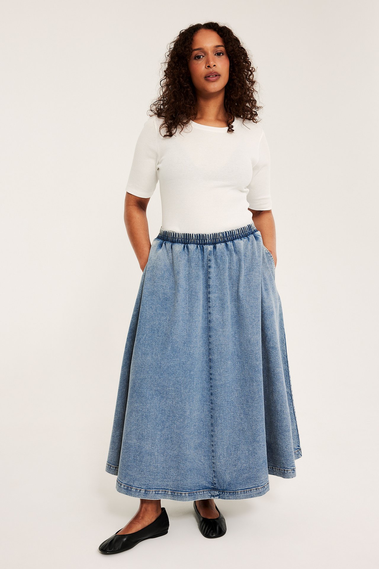 Framifrån: Kvinna i ljusblå midilång jeanskjol med resår i midjan, vit topp och svarta ballerinaskor.