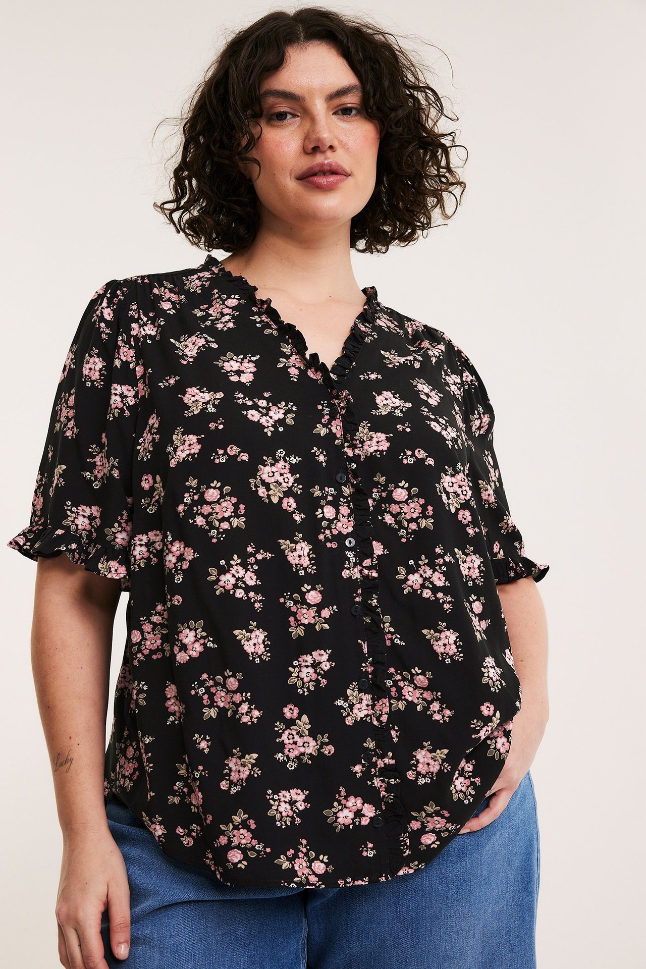Front view: kvinna i svart blommig viskosblus med volanger, klädda knappar och blå jeans.