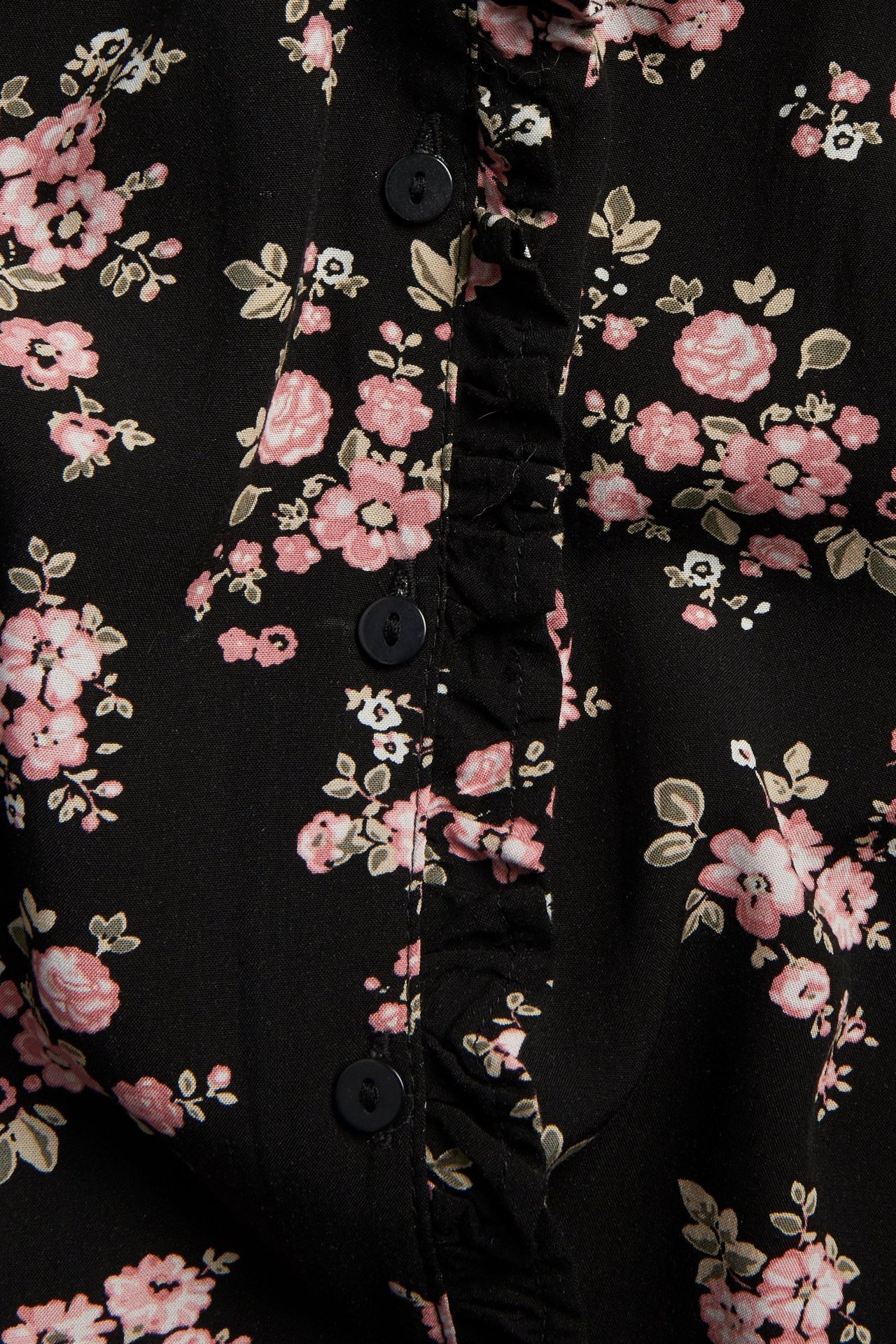 Nærbilde av svart viskosebluse med rosa blomstermønster, rysjedetalj og svarte knapper.