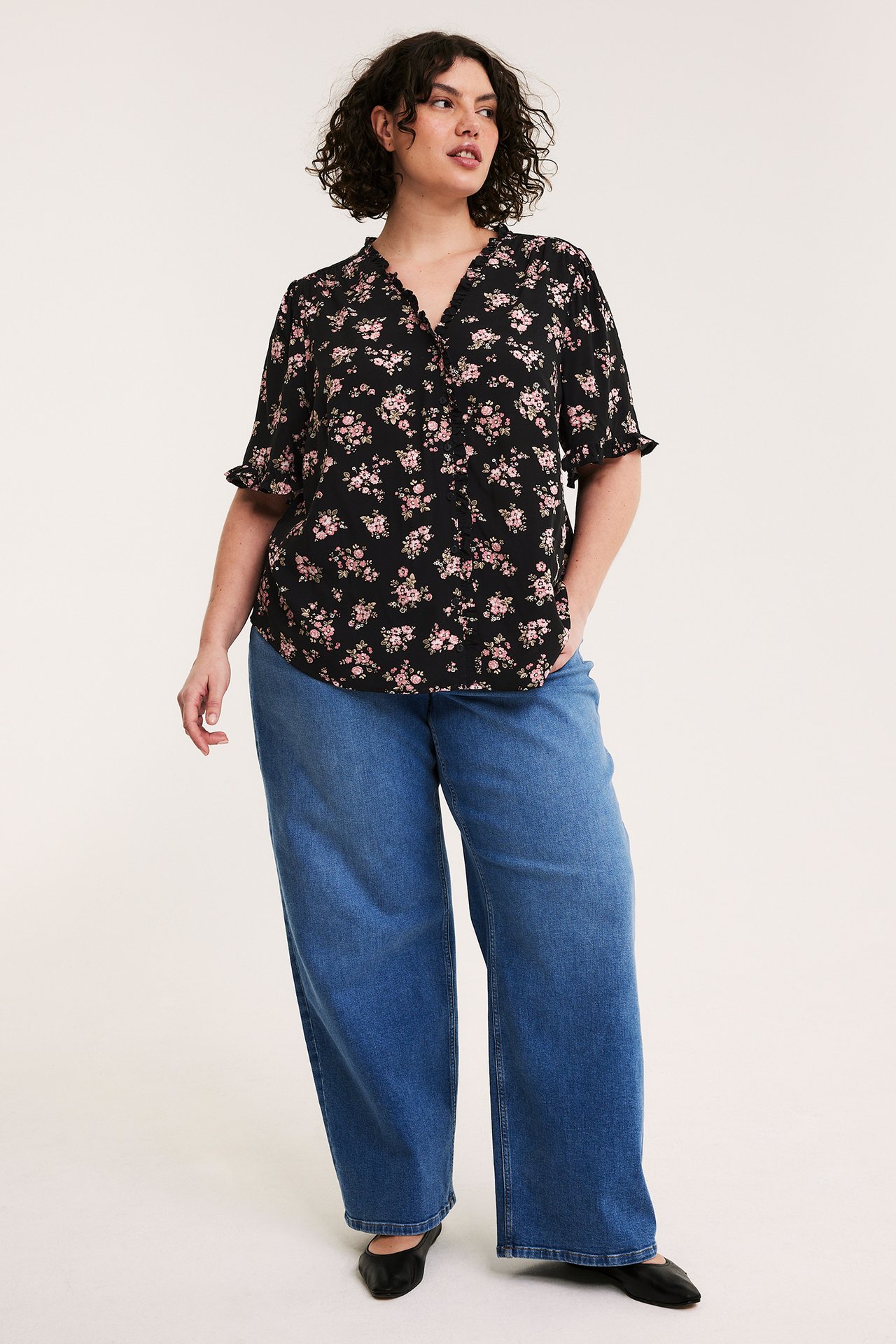 Framsida av kvinna i svart blommig V-ringad blus med volanger, blå vida jeans och svarta platta skor.