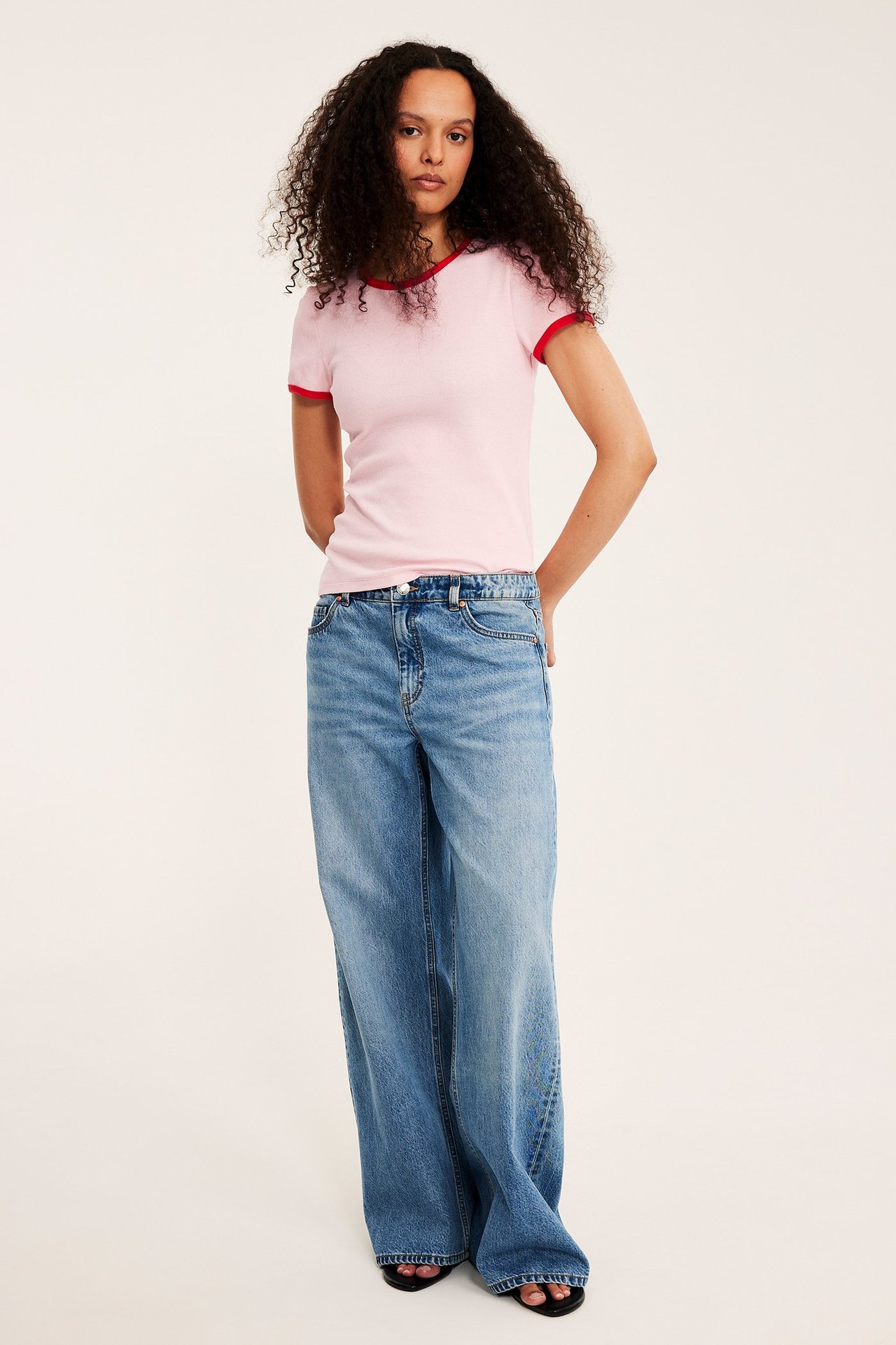 Edestäpäin nainen: vaaleanpunainen ribattu toppi punaisilla reunoilla, siniset leveät farkut, mustat sandaalit.