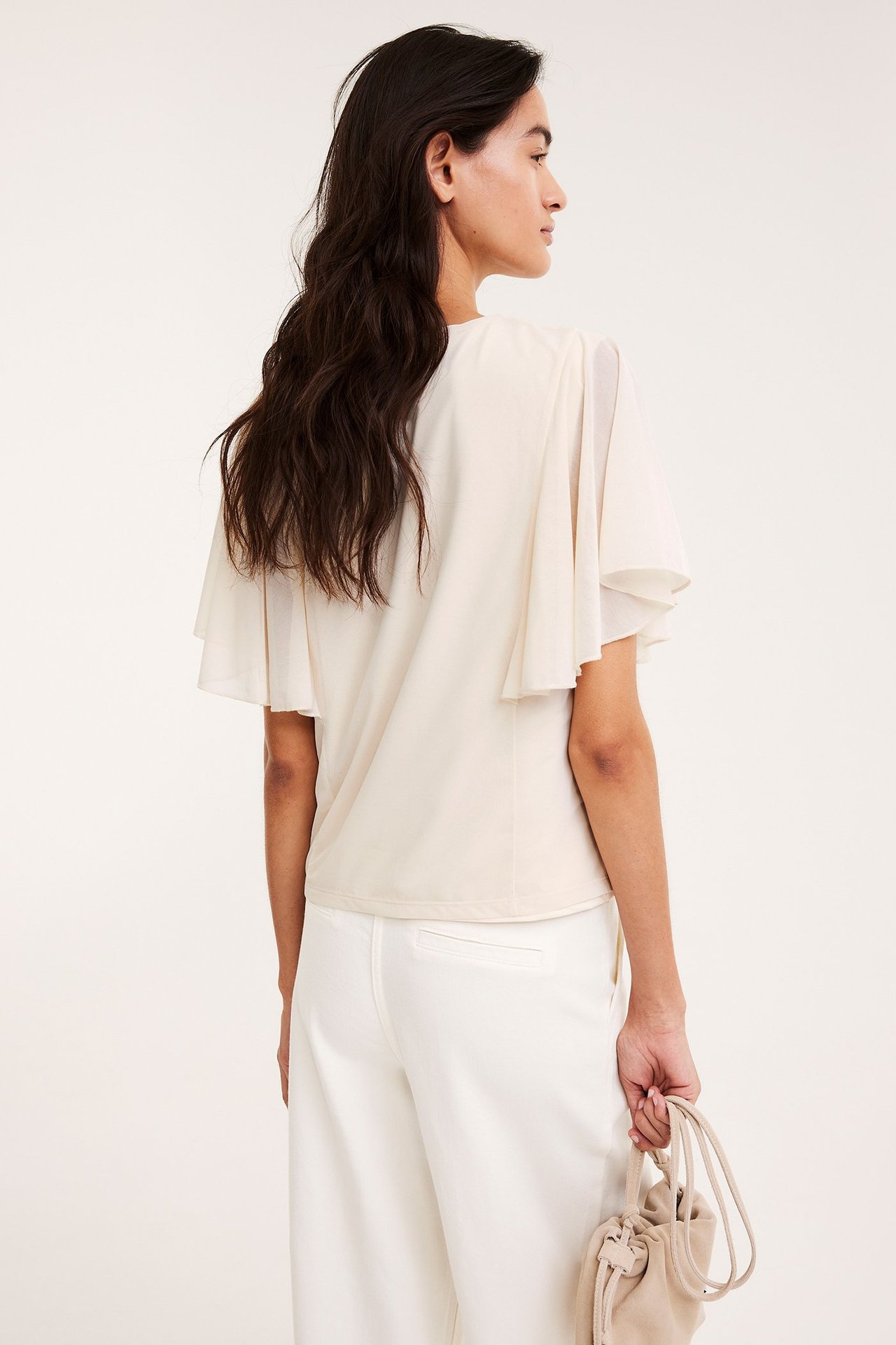 Bak-/sidovy av kvinna i off-white skir topp med vida ängelsärmar, vita byxor, hållandes en beige väska.