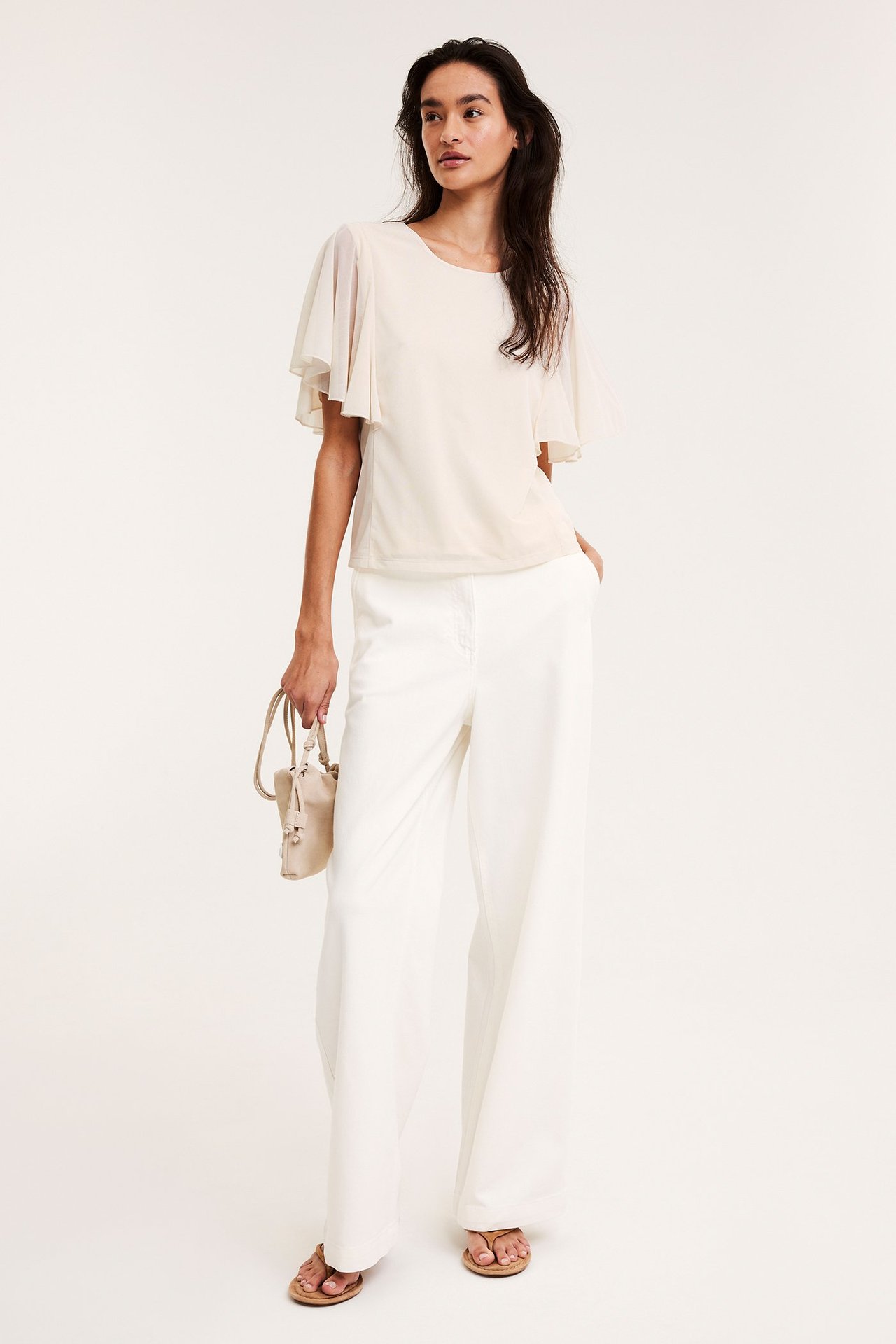 Kvinne i offwhite gjennomsiktig topp med engleermer, hvite bukser, beige sandaler, holder beige veske.