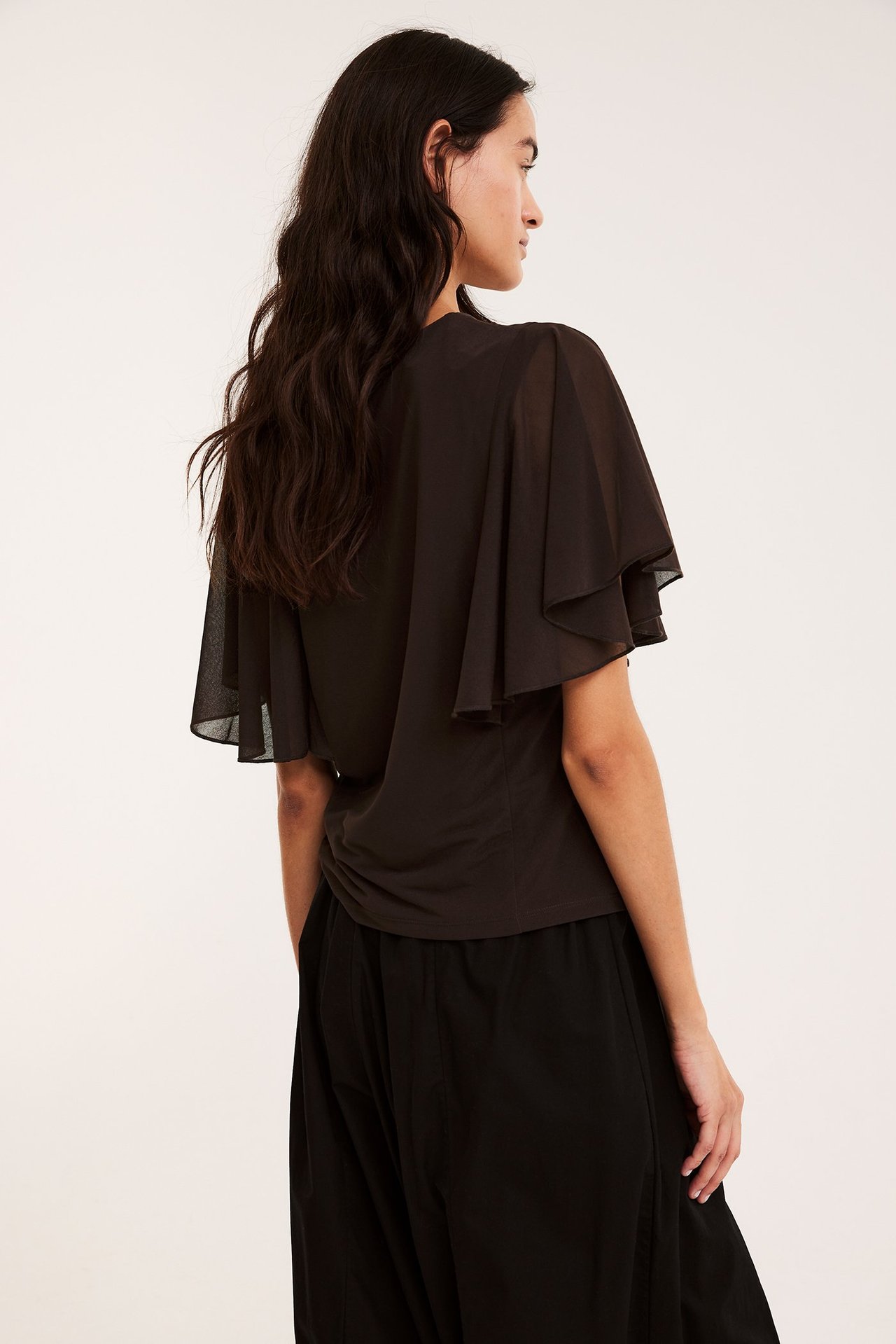 Baksidesvy av kvinna i mörkbrun skir topp med korta vida ärmar och svarta byxor.