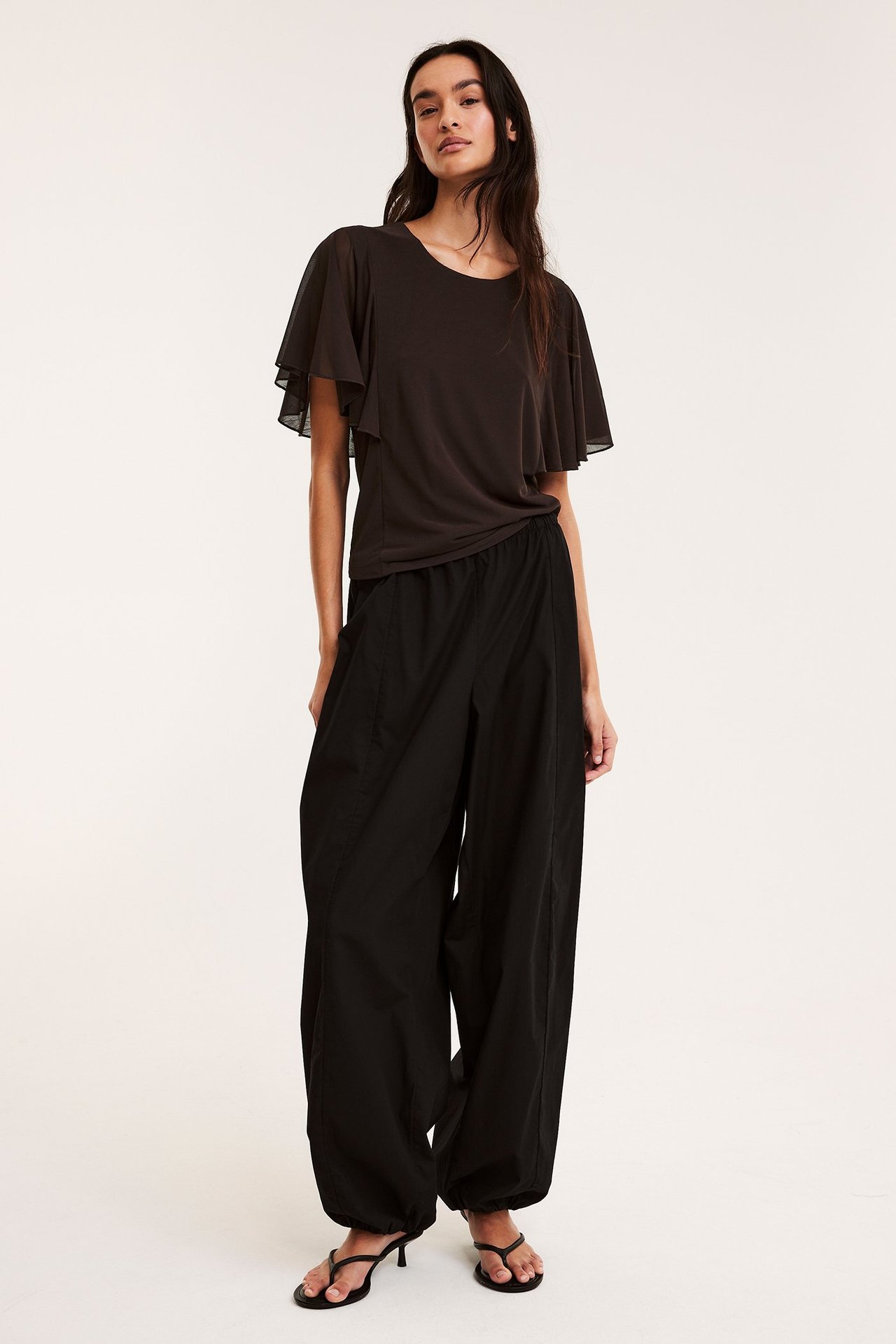 Framifrån, kvinna i mörkbrun skir topp med korta vida ärmar, svarta vida byxor och svarta klacksandaler.