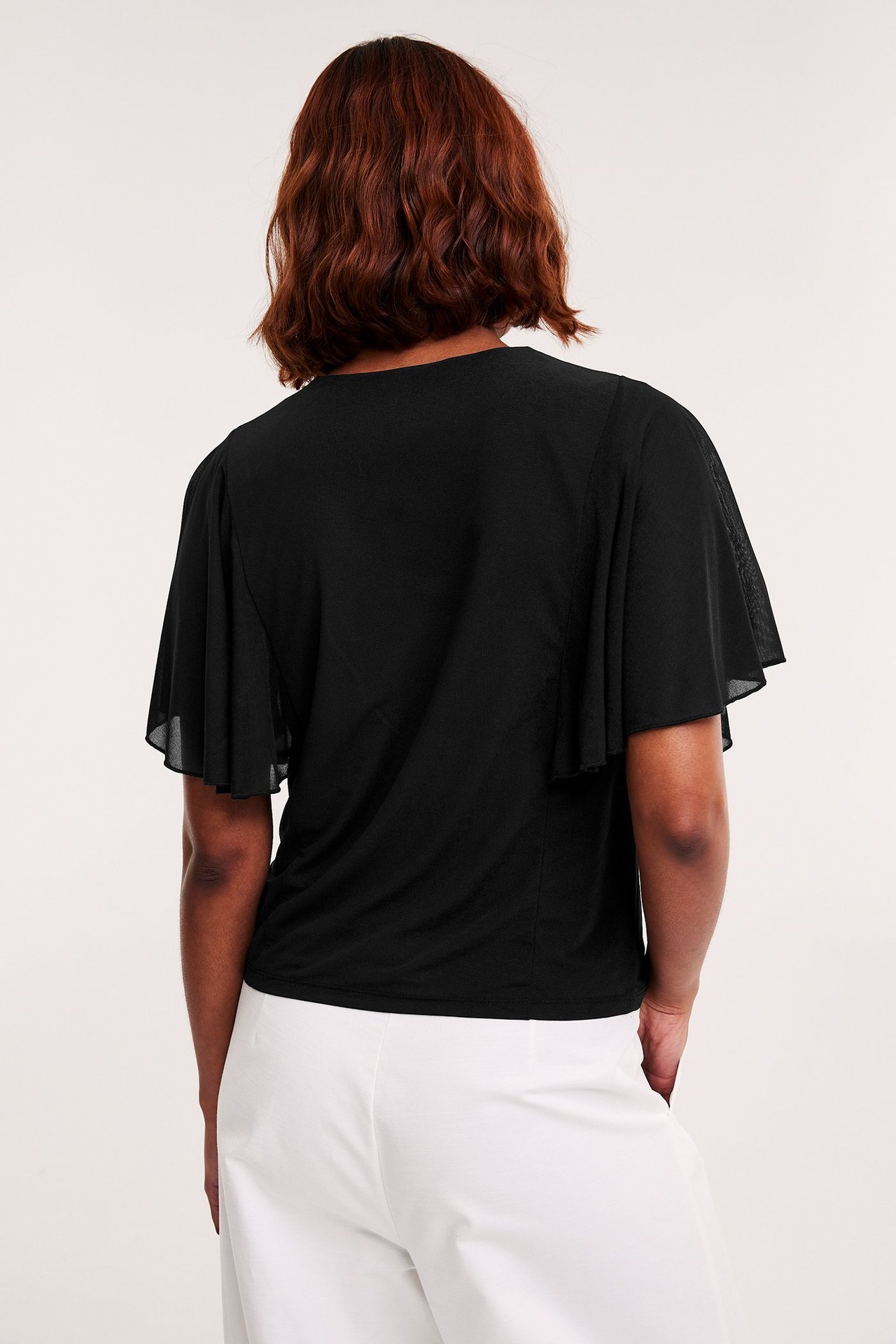 Baksida av kvinna i svart transparent topp med vida ängelsärmar och vita vida byxor.