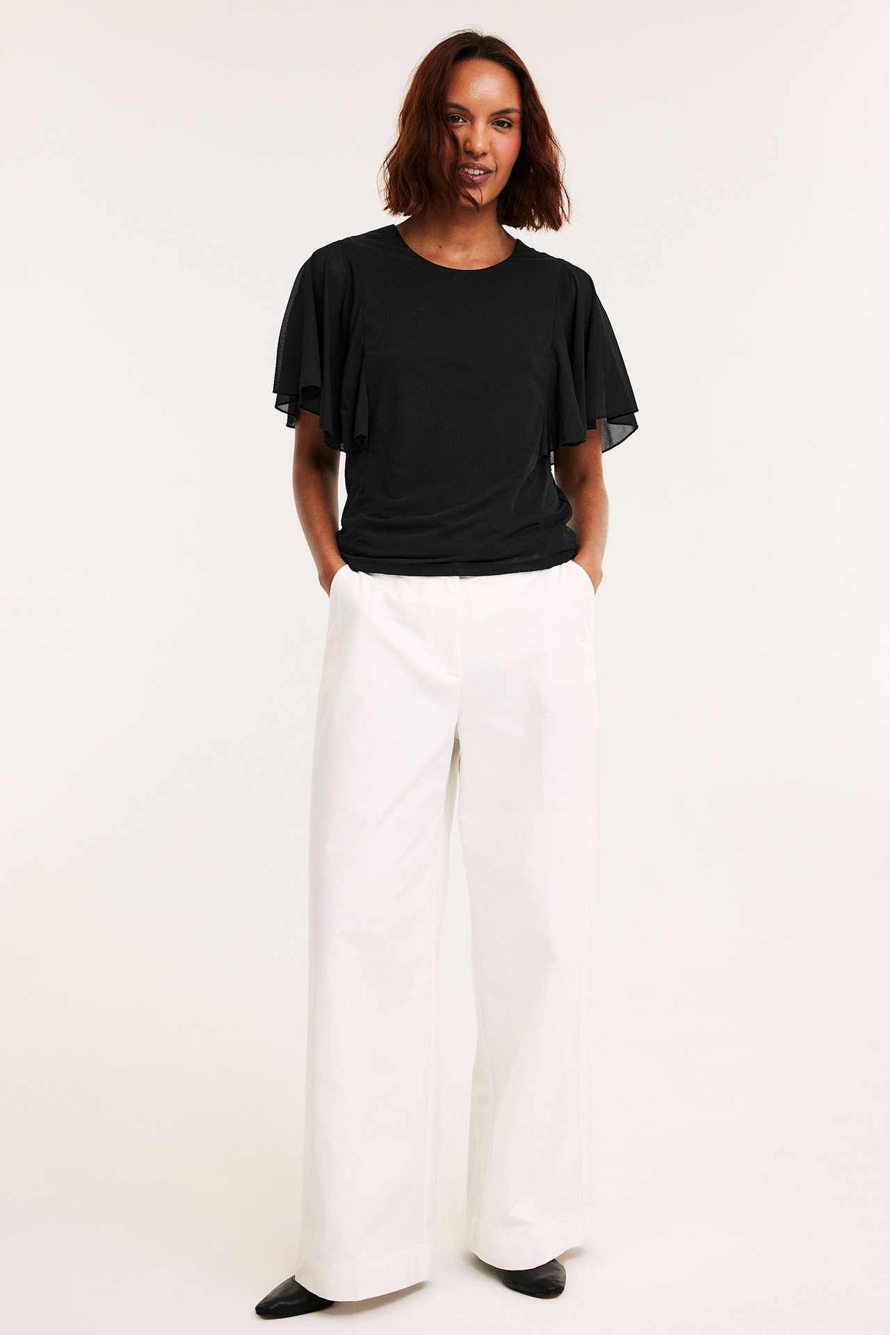 Framifrån: kvinna i svart transparent topp med vida ärmar, vita vida byxor och svarta skor.