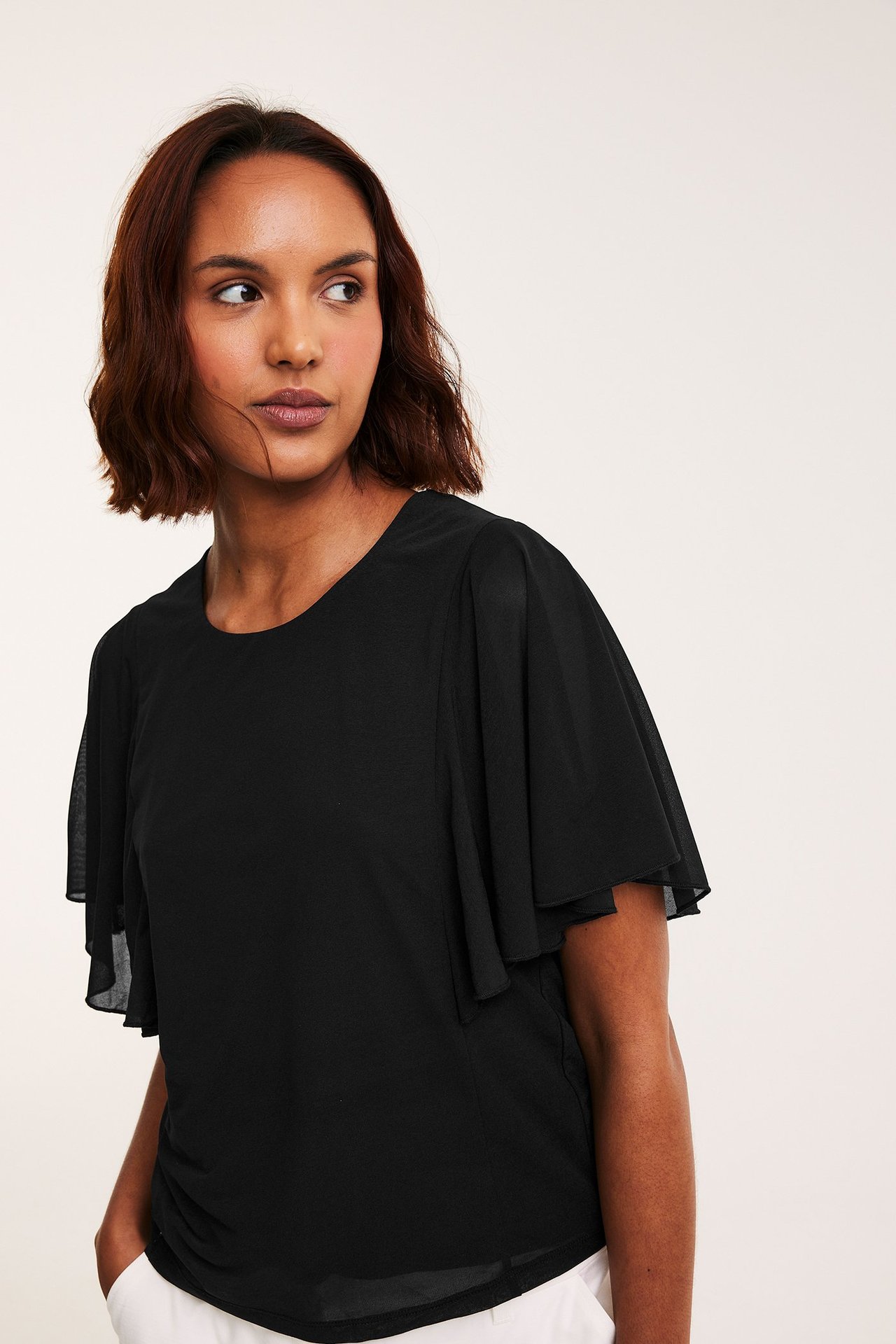 Framifrån, kvinna i svart transparent topp med korta vida änglaärmar, stylad med vita byxor.