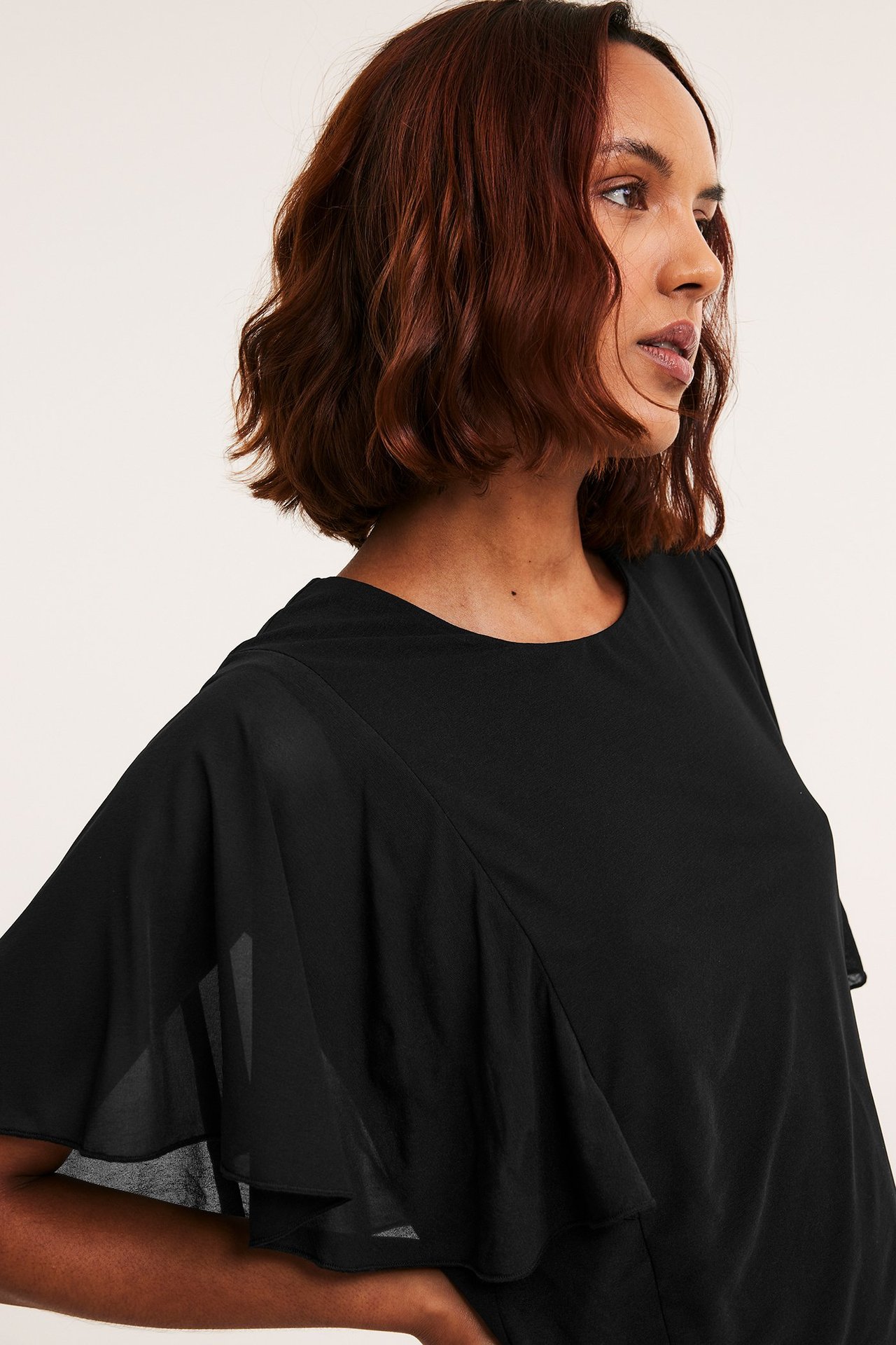Sidebilde av en kvinne i en løstsittende svart gjennomsiktig topp med volangermer og rund hals.