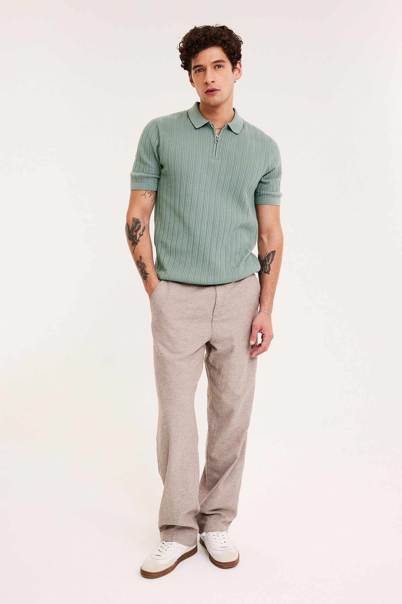 Edestä: mies vaaleanvihreässä ribbineulotussa vetoketjullisessa pikeepaidassa, beigessä housuissa ja valkoisissa tennareissa.