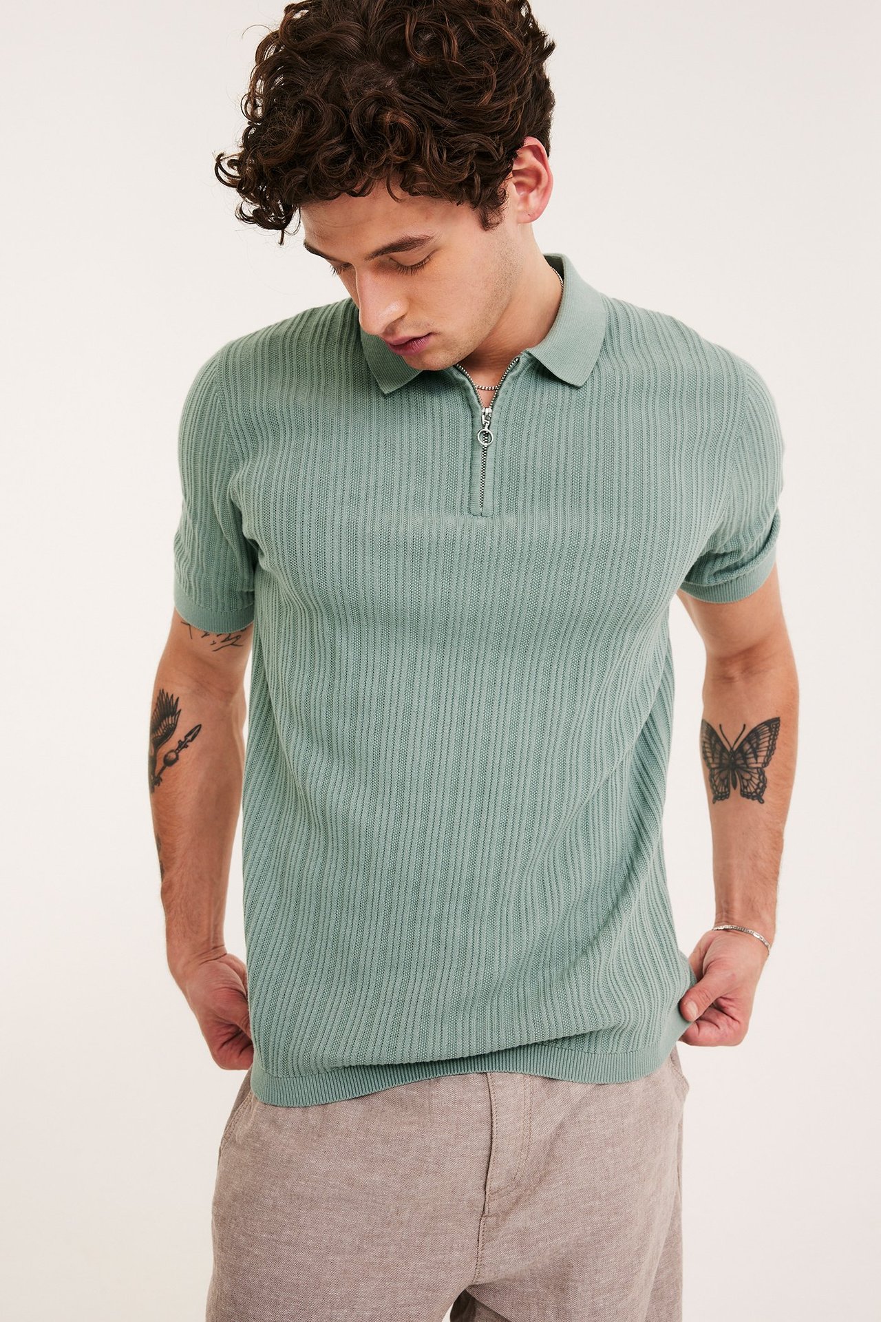 Framifrån: man i ljusgrön ribbstickad pikétröja med silver half-zip, stylad med beige shorts.