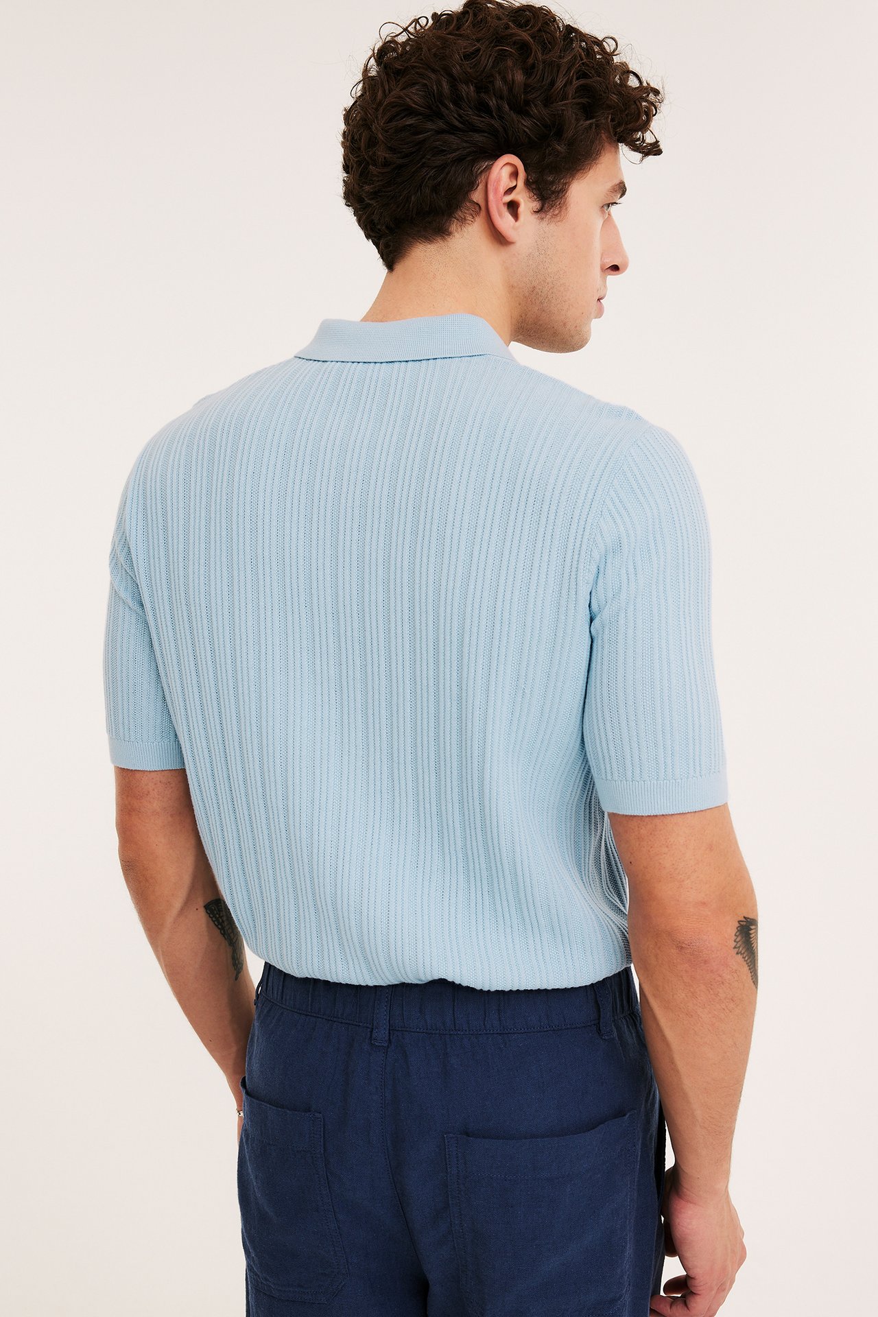 Widok z tyłu na mężczyznę w jasnoniebieskiej prążkowanej dzianinowej koszulce polo i granatowych szortach.