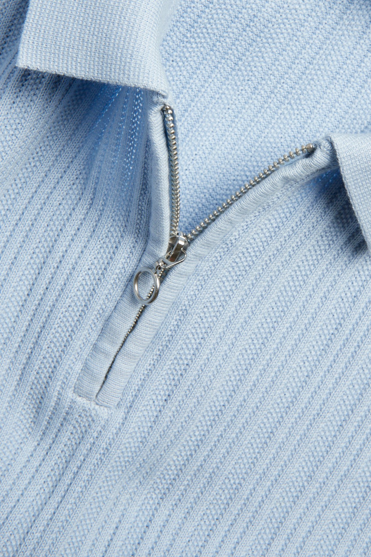 Nærbilde av en lyseblå strikket piké med sølvfarget half-zip og strukturert mønster.