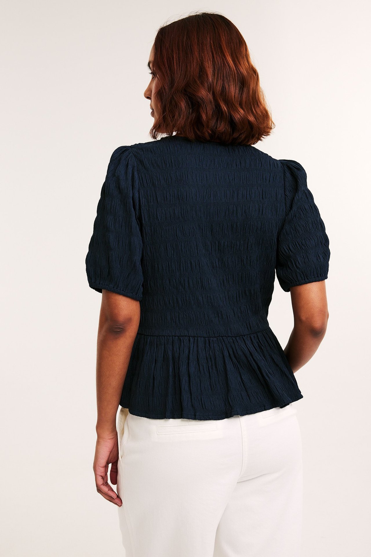 Naisen selkä, yllään tummansininen, kuvioitu, pöyhkeähihainen peplumtoppi ja vaaleat housut.