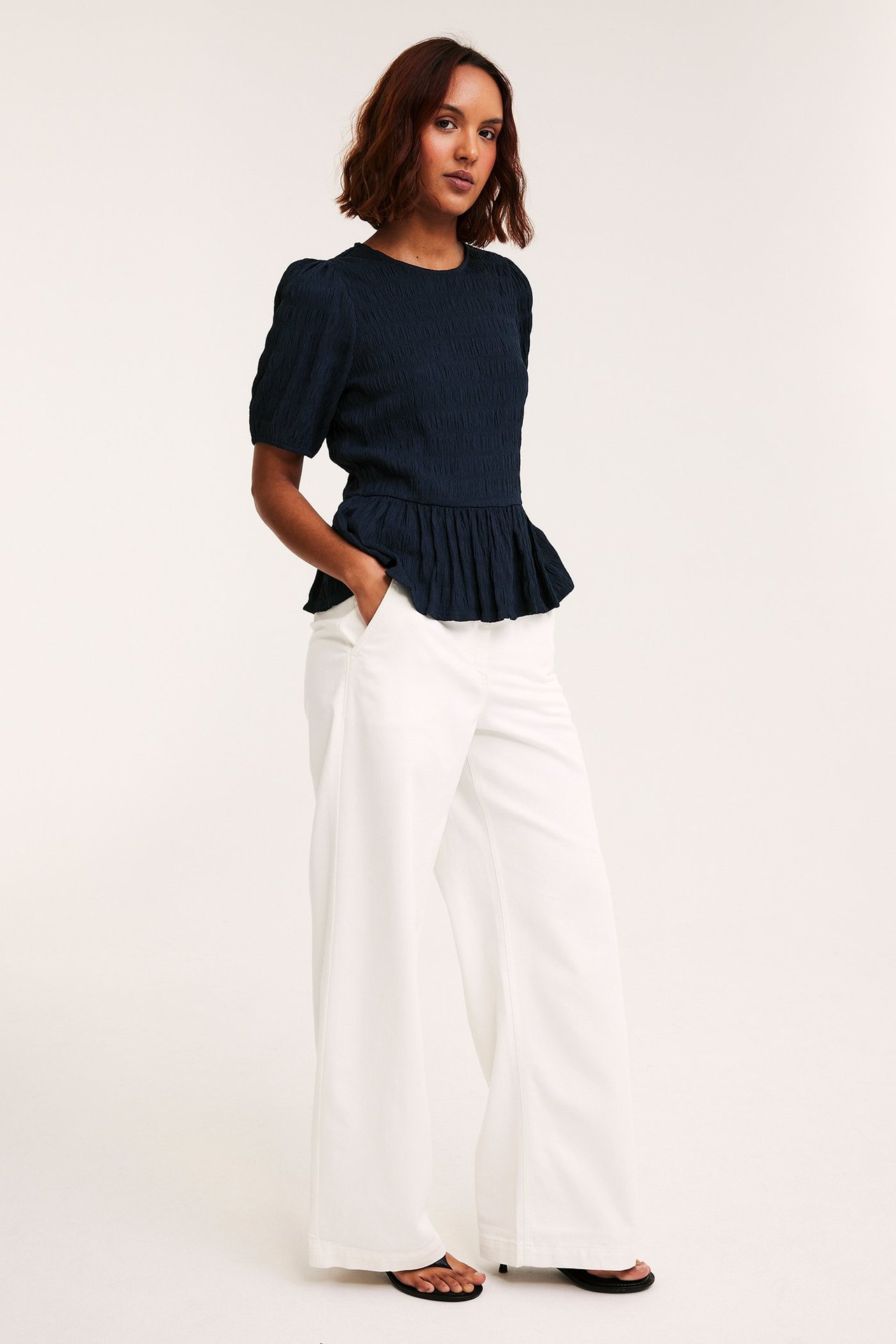 Frontbilde: kvinne i mørkeblå strukturert peplumtopp med puffermer og hvite vide bukser.