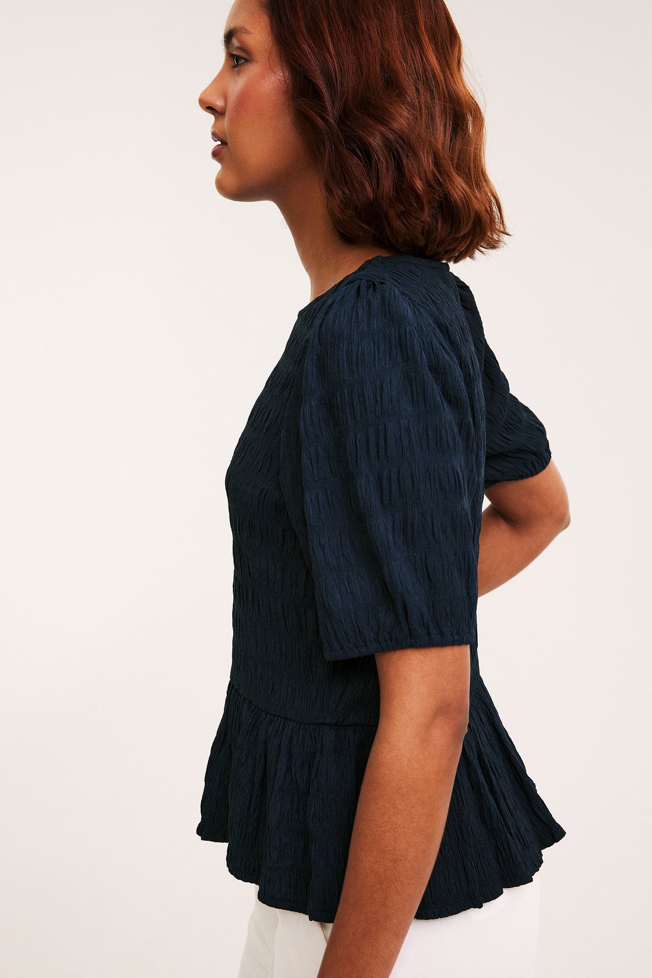 Naisen sivukuva tummansinisessä strukturoidussa peplum-topissa, jossa pussiholkit, sekä valkoisissa housuissa.
