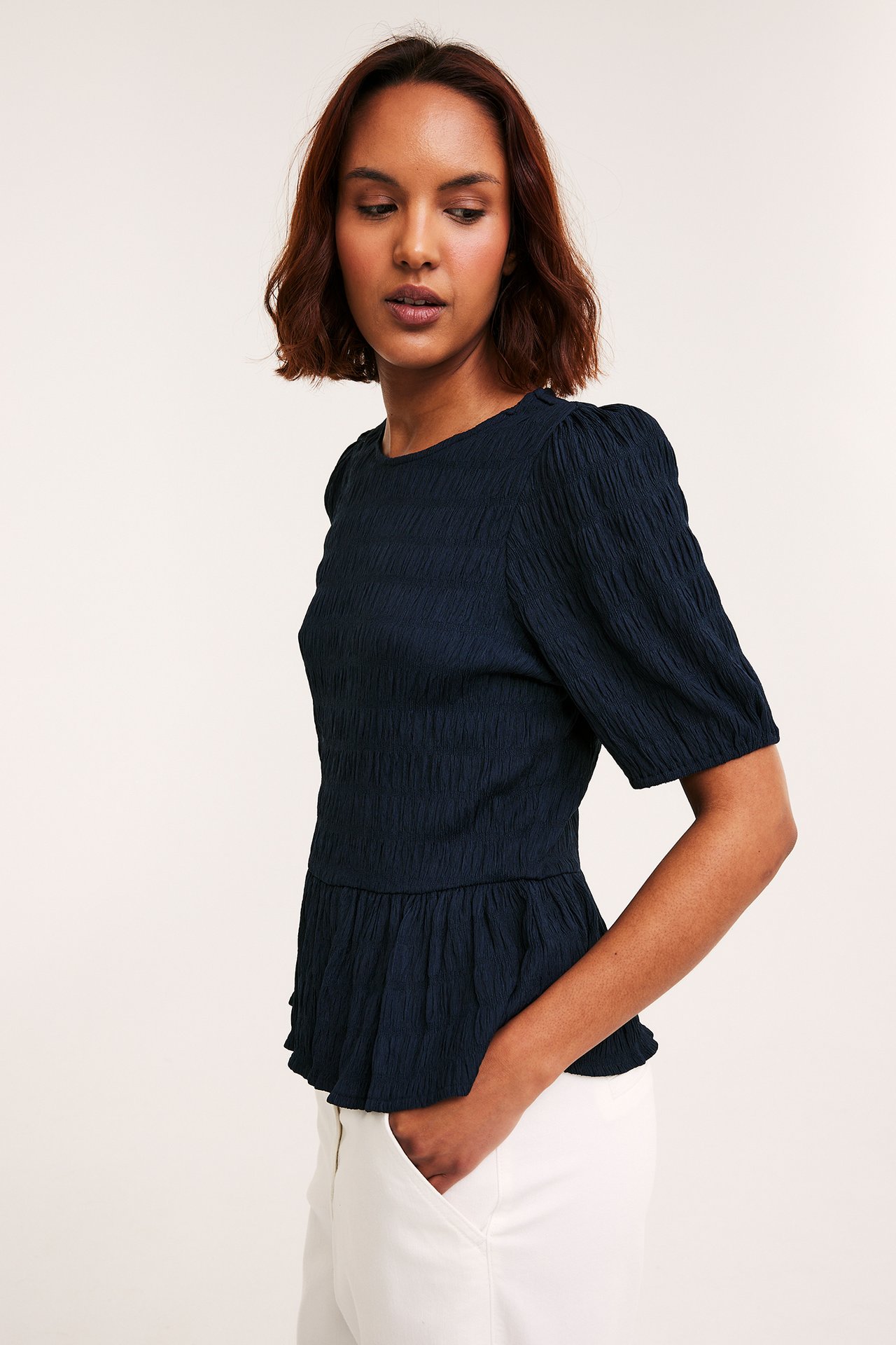 Naisen etunäkymä tummansinisessä strukturoidussa peplumtopissa puffihihoilla ja valkoisissa housuissa.