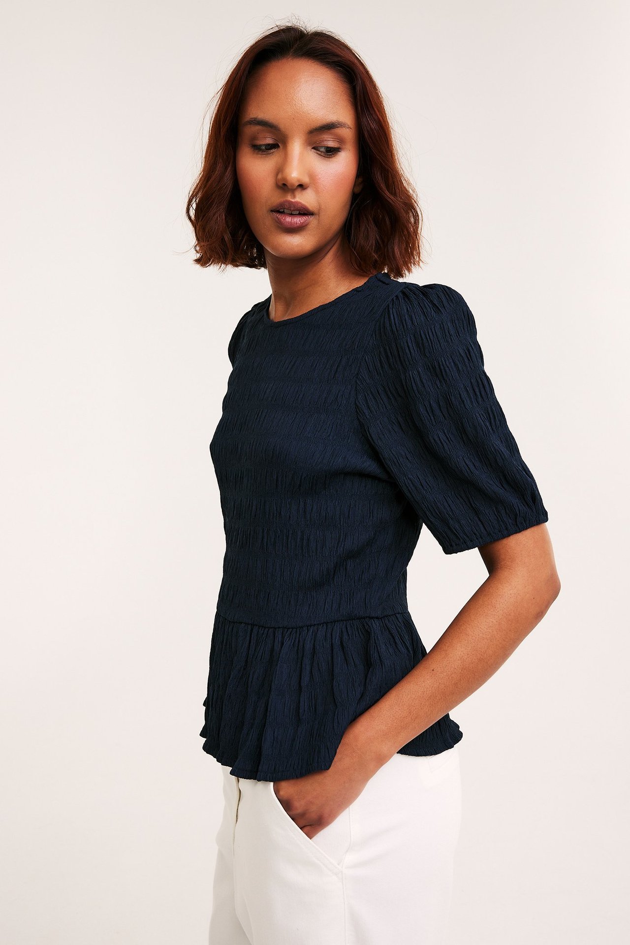 Frontbilde av en kvinne i mørkeblå, strukturert peplumtopp med puffermer og hvite bukser.