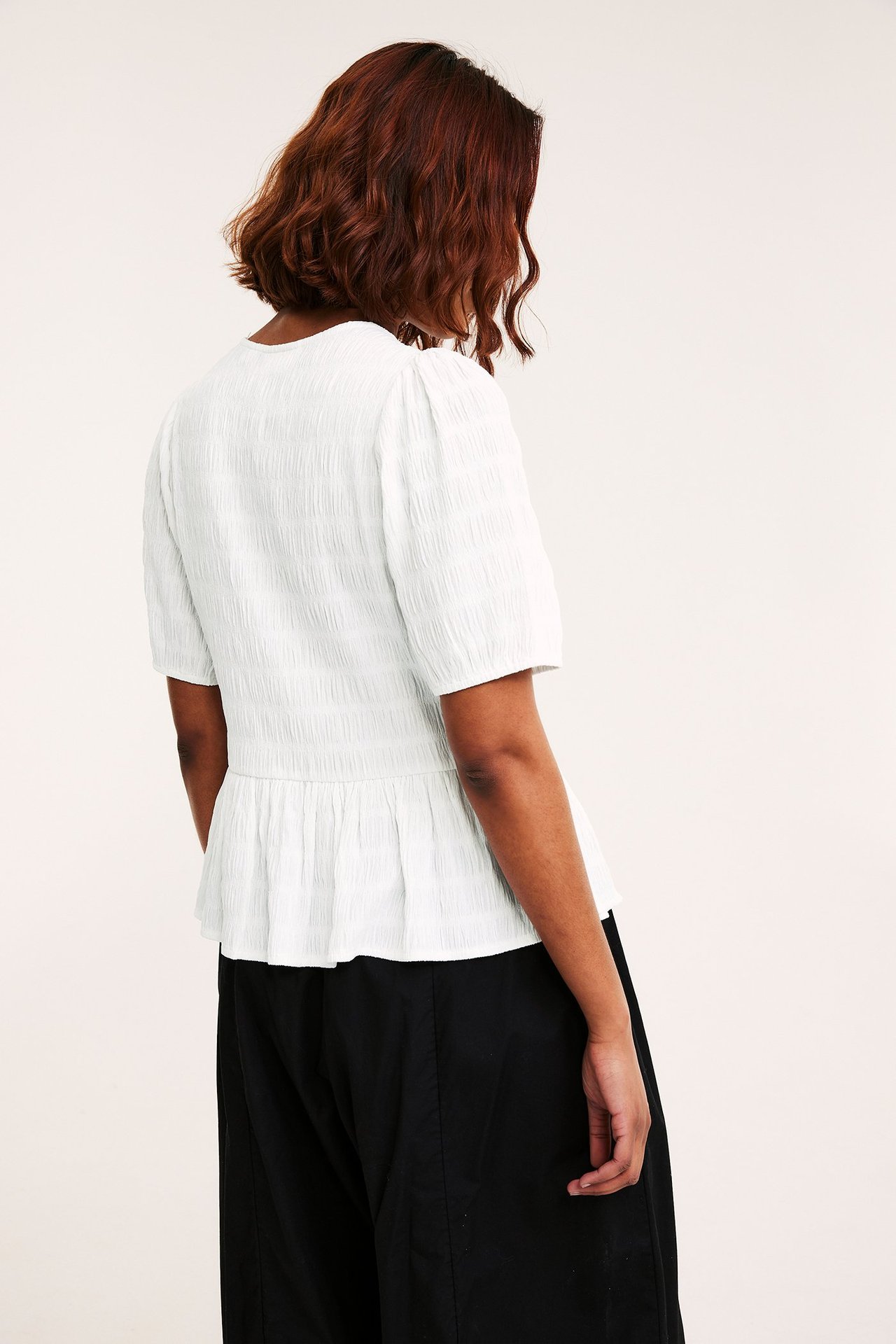 Ryggvisning av kvinne i offwhite strukturert peplumtopp med puffermer og svarte vide bukser.