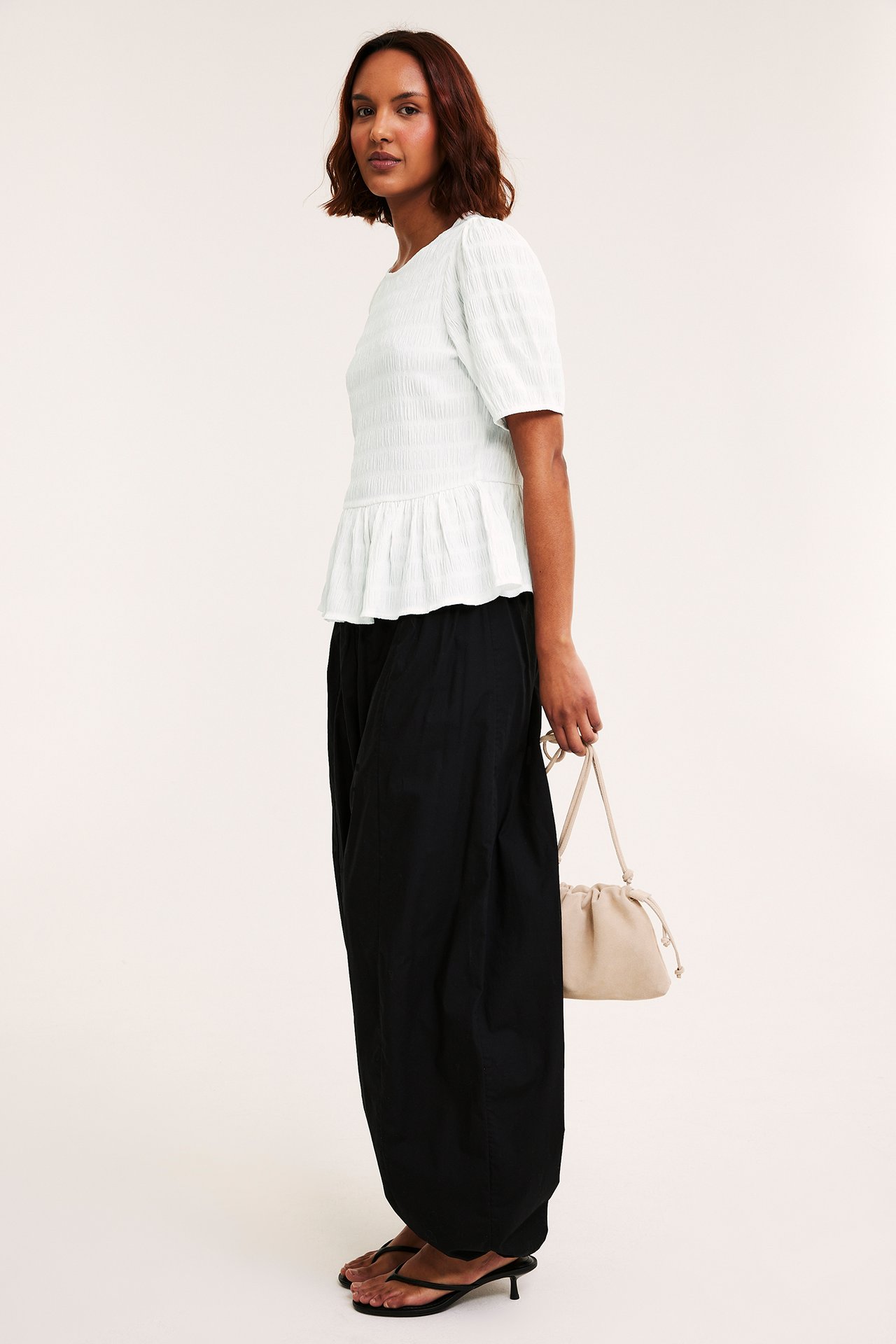 Sidebilde: Kvinne i offwhite strukturert peplumtopp, svarte vide bukser, svarte sandaler, holder beige snøreveske.