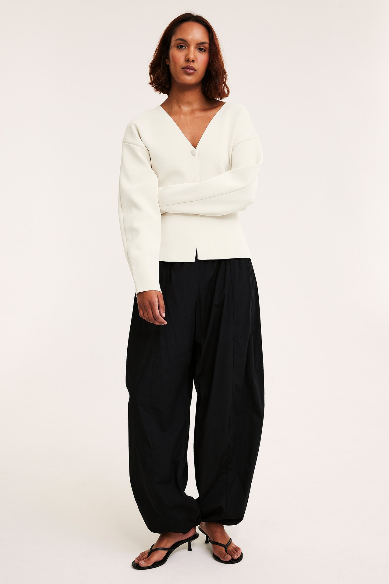 Edestä: Nainen yllään luonnonvalkoinen V-kauluksinen neuletakki, mustat löysät housut ja mustat korkeakorkoiset sandaalit.