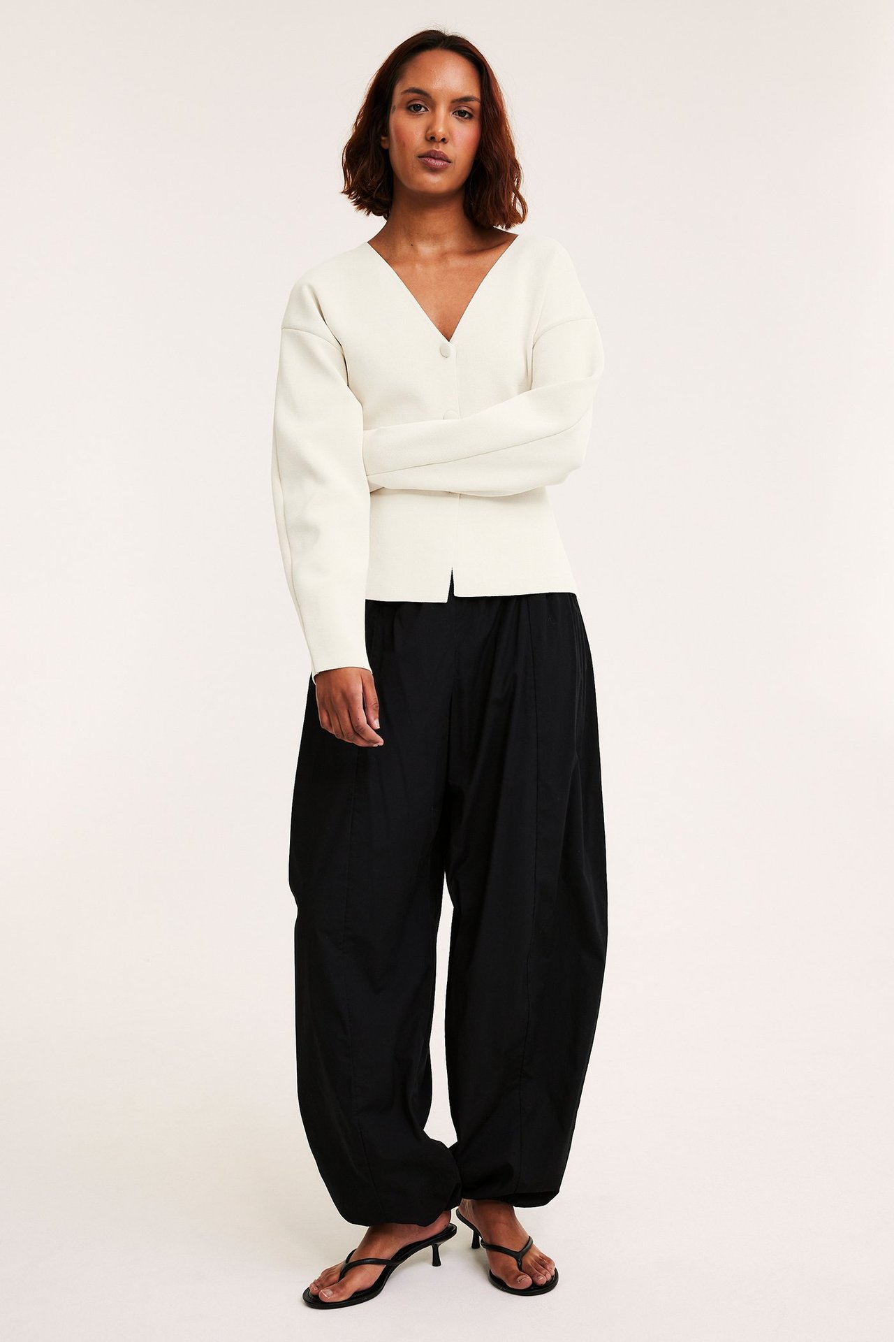 Kobieta w białym, taliowanym kardiganie z dekoltem V, luźnych czarnych spodniach i czarnych sandałach na obcasie.