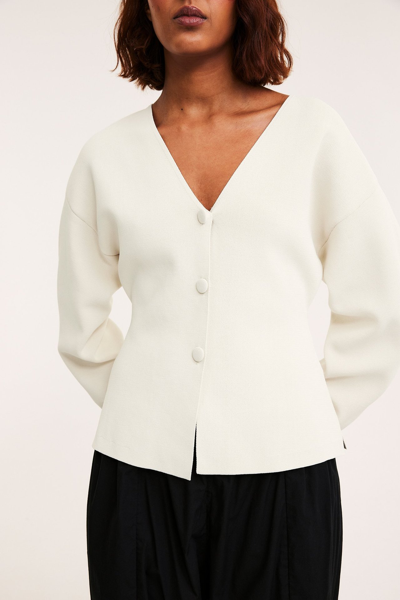 Framifrån, kvinna i en offwhite V-ringad cardigan med insvängd midja, tre knappar, över svarta byxor.