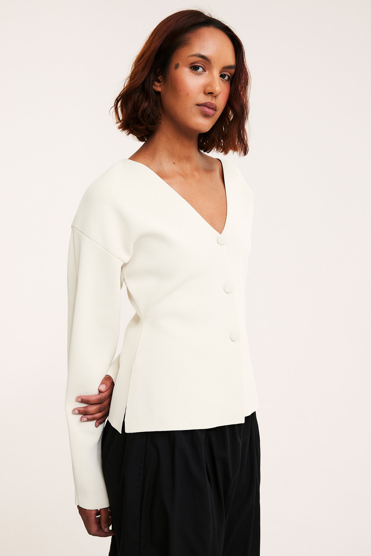 Edestä: Nainen offwhite polyesteri V-kauluksellisessa neuletakissa kolmella napilla ja mustissa housuissa.