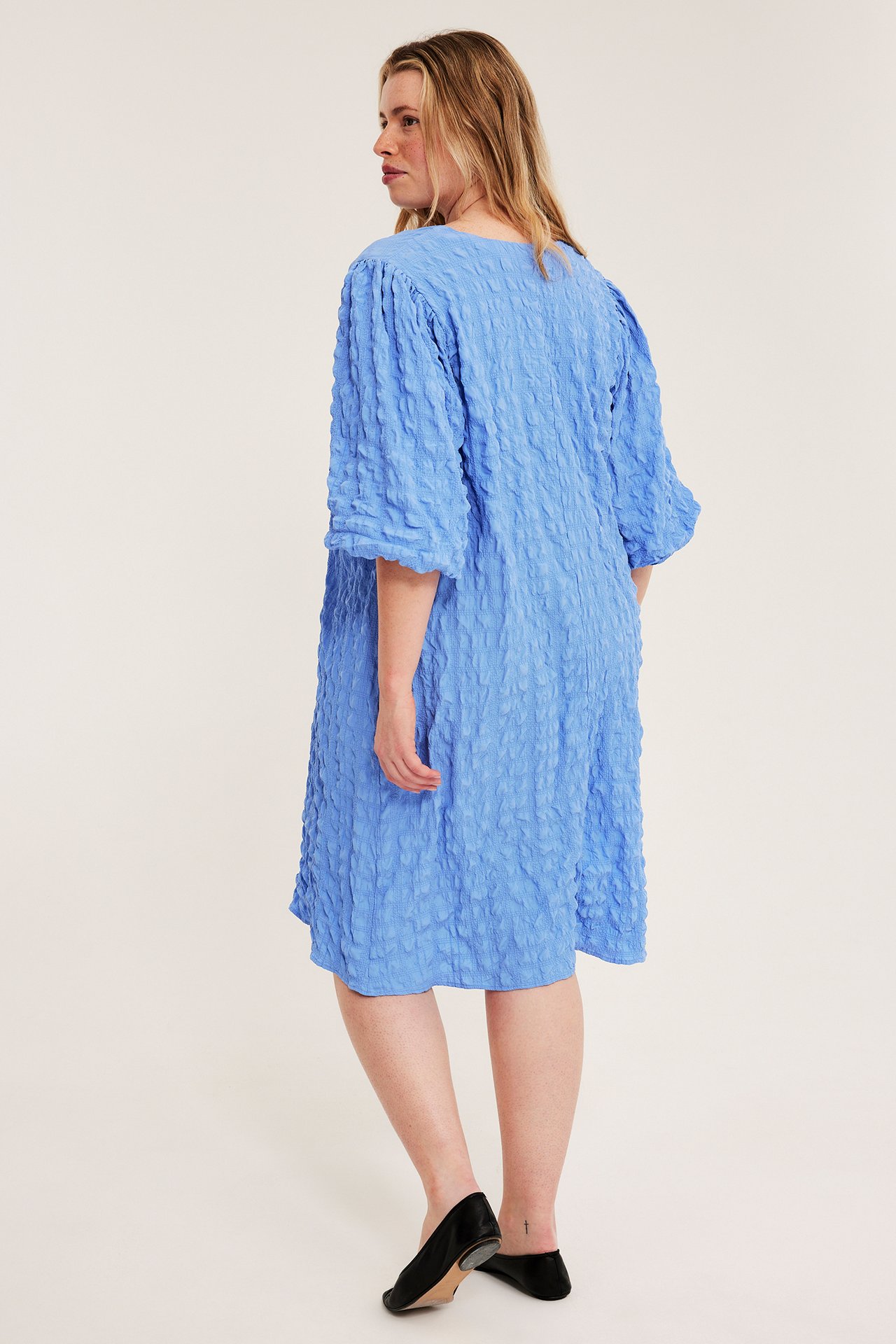 Widok z boku: kobieta w niebieskiej sukience midi z dekoltem V, bufiastymi rękawami i czarnymi baletkami.