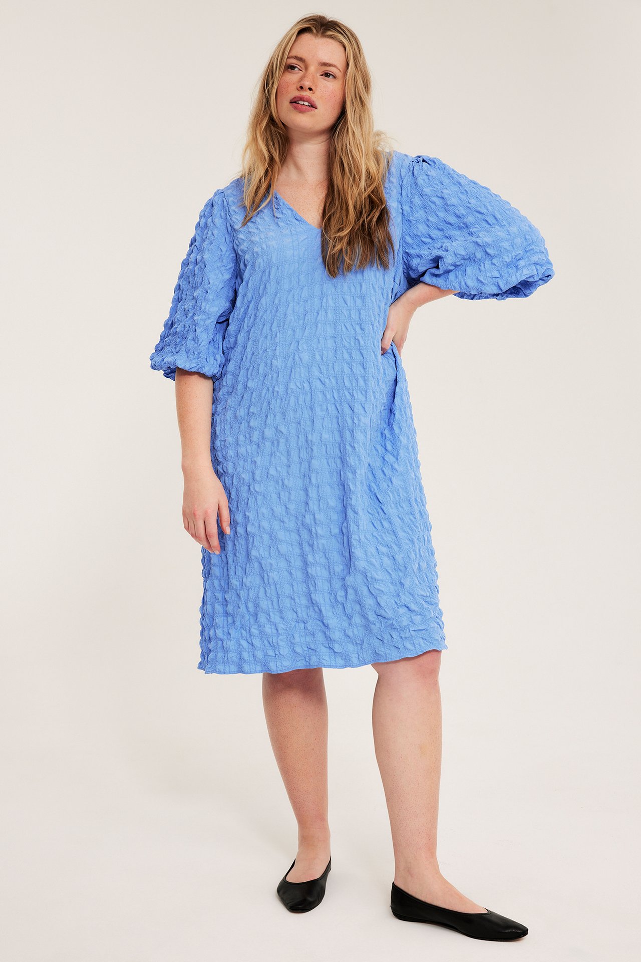 Frontvy av en kvinna i en blå knälång strukturerad V-ringad klänning med puffärmar och svarta platta skor.