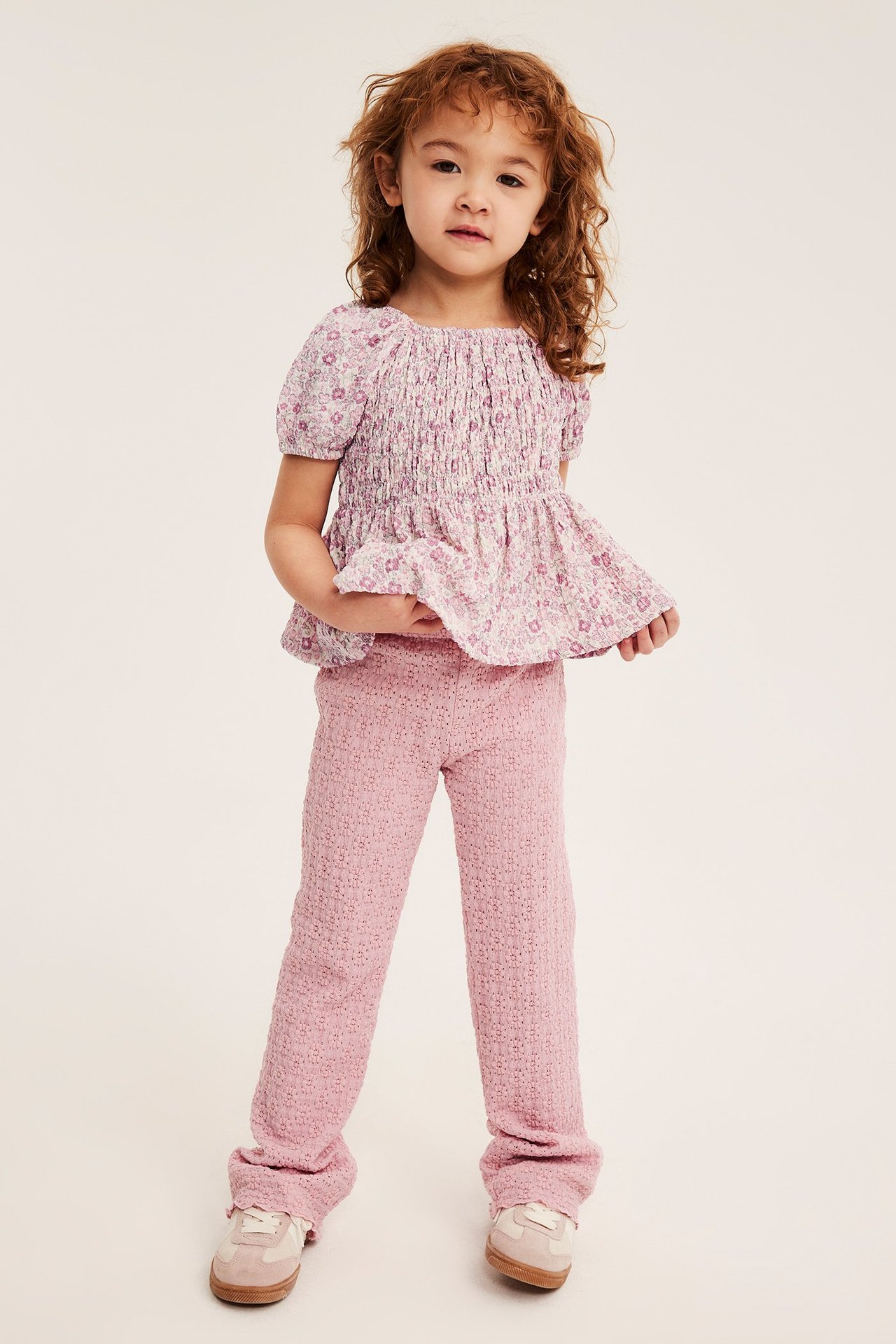 Framifrån: Ett barn i offwhite blommig smocktopp, rosa texturerade byxor och beiga sneakers.