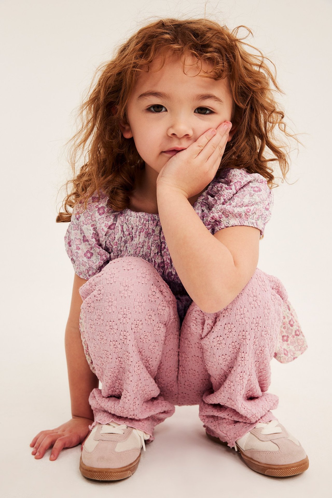 Frontvy av ett barn i offwhite blommig smocktopp, rosa texturerade byxor och beige sneakers.