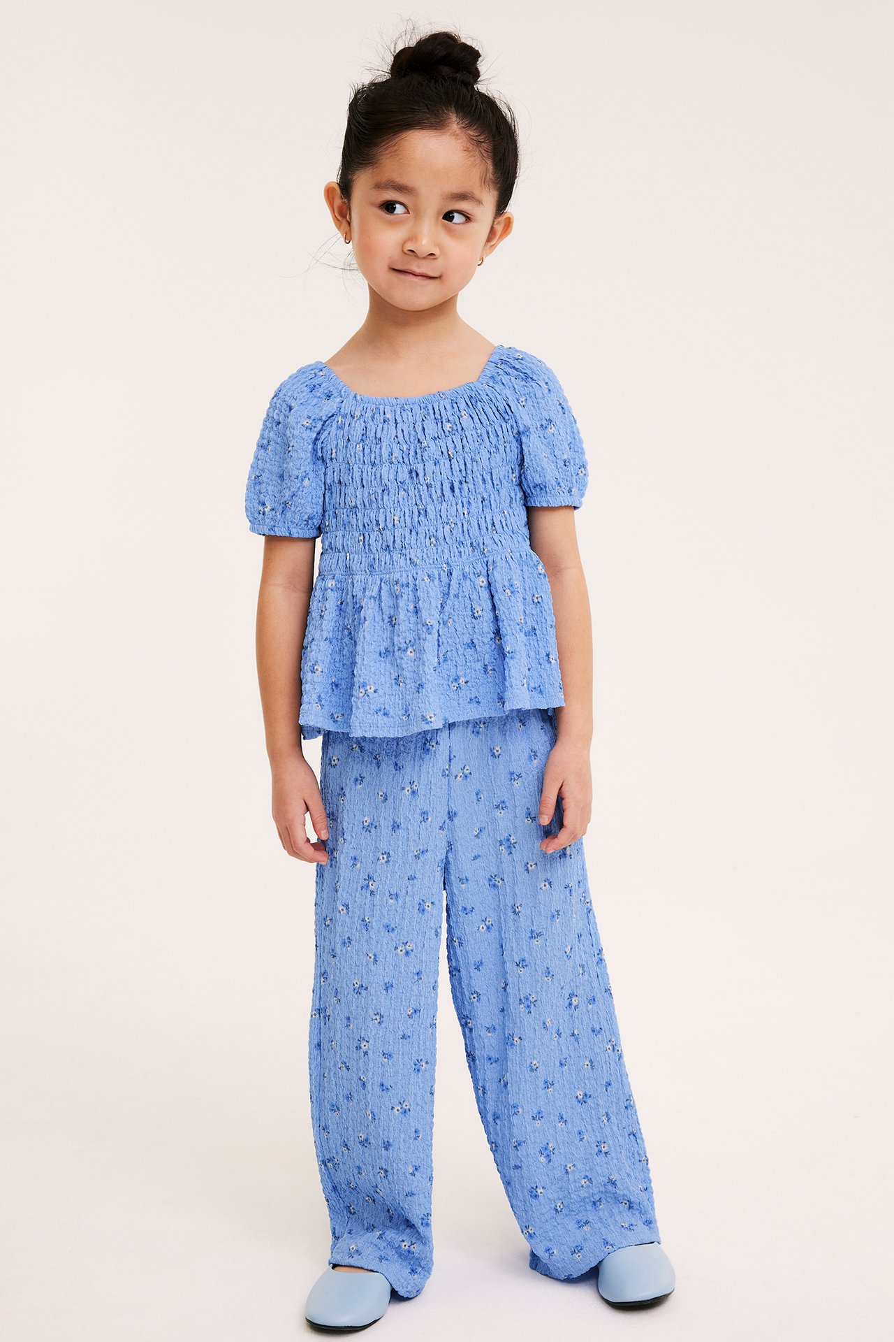 Frontvy av ett barn som bär en ljusblå blommig smocktopp med puffärmar, matchande vida byxor och ljusblå ballerinaskor.