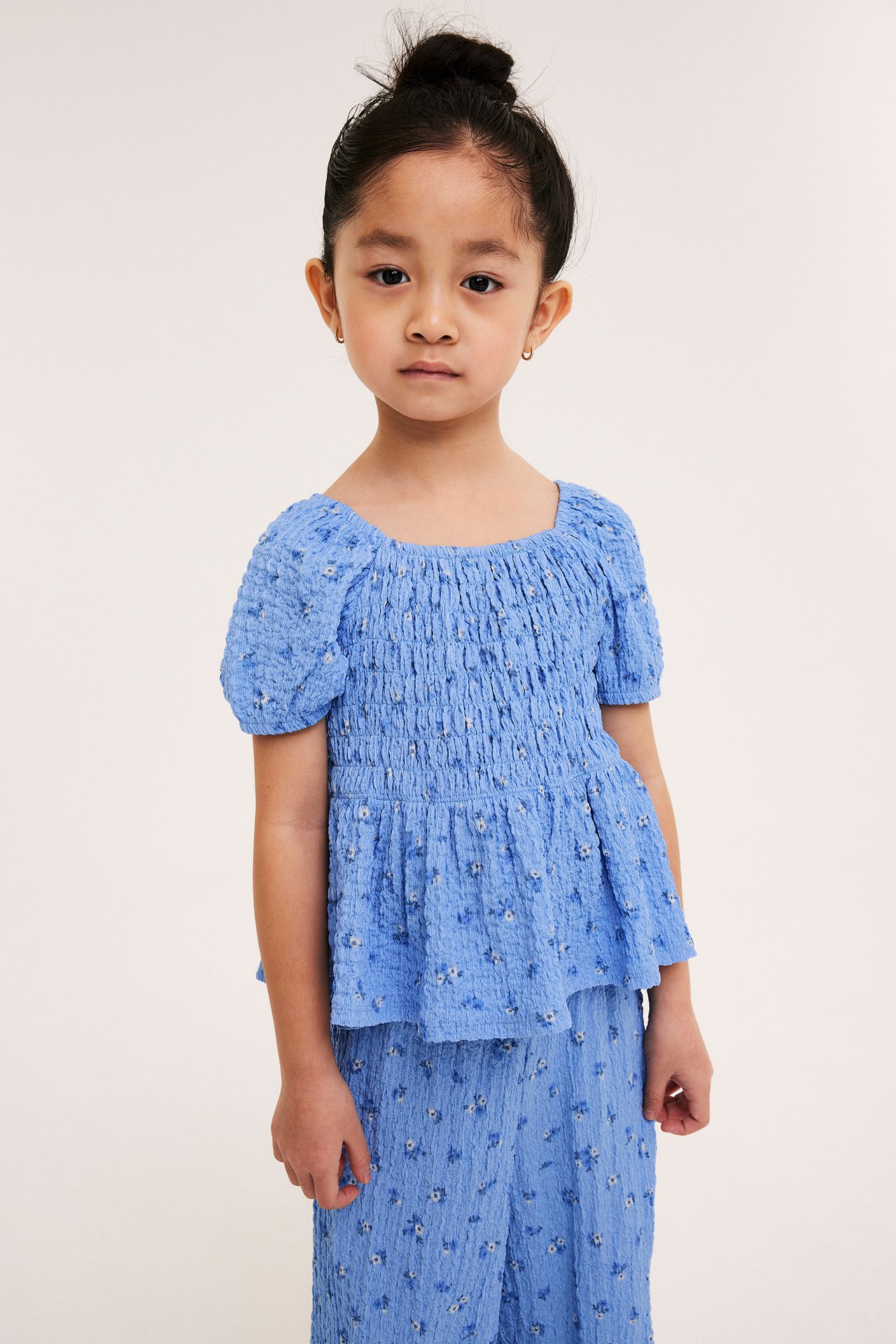 Framsida av ett barn i en ljusblå smockad blommig topp med korta puffärmar och matchande byxor.