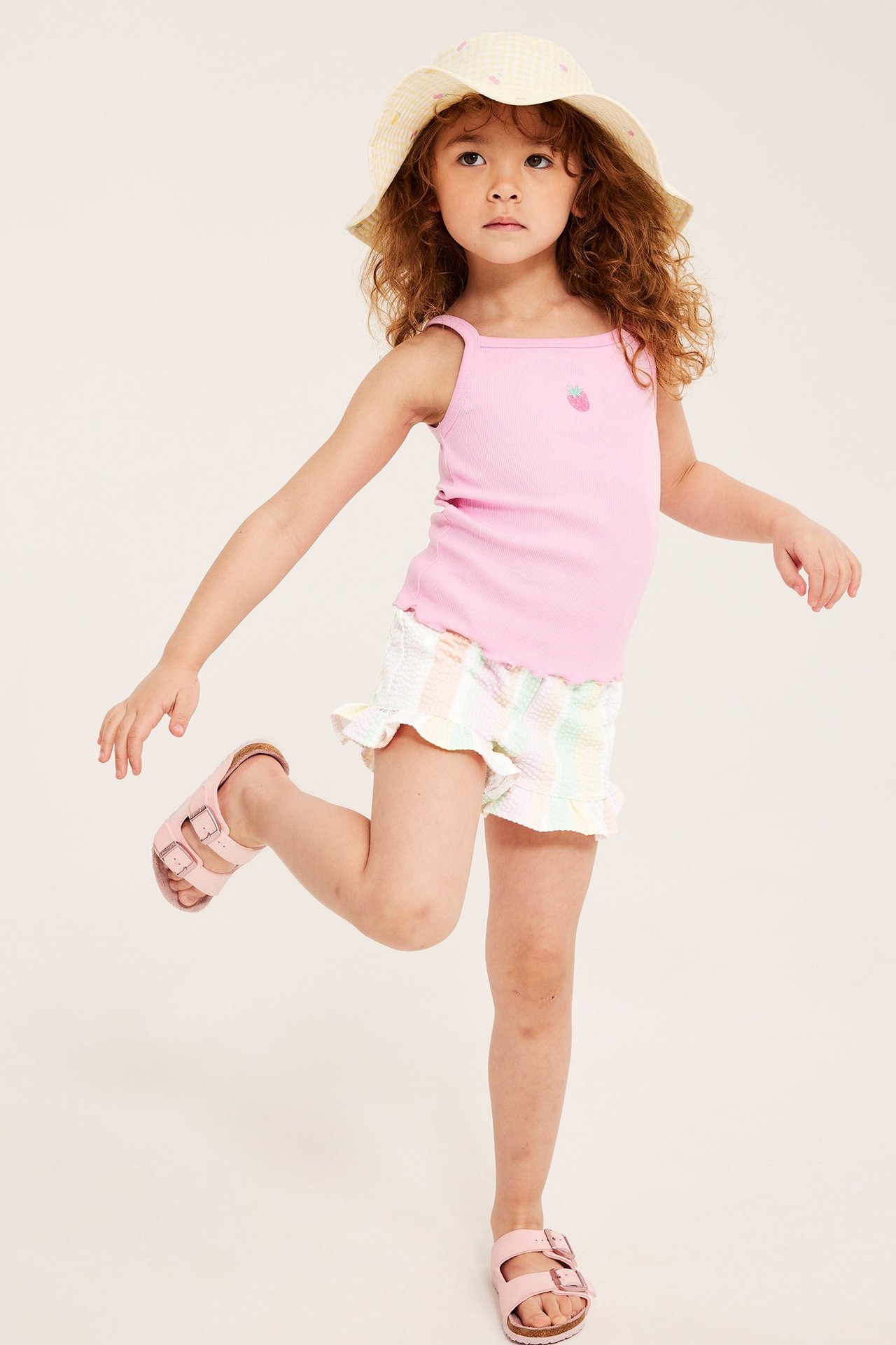 Lapsi yllään luonnonvalkoiset pastelliraitaiset shortsit röyhelöillä, pinkki toppi, keltainen hattu ja pinkit sandaalit.