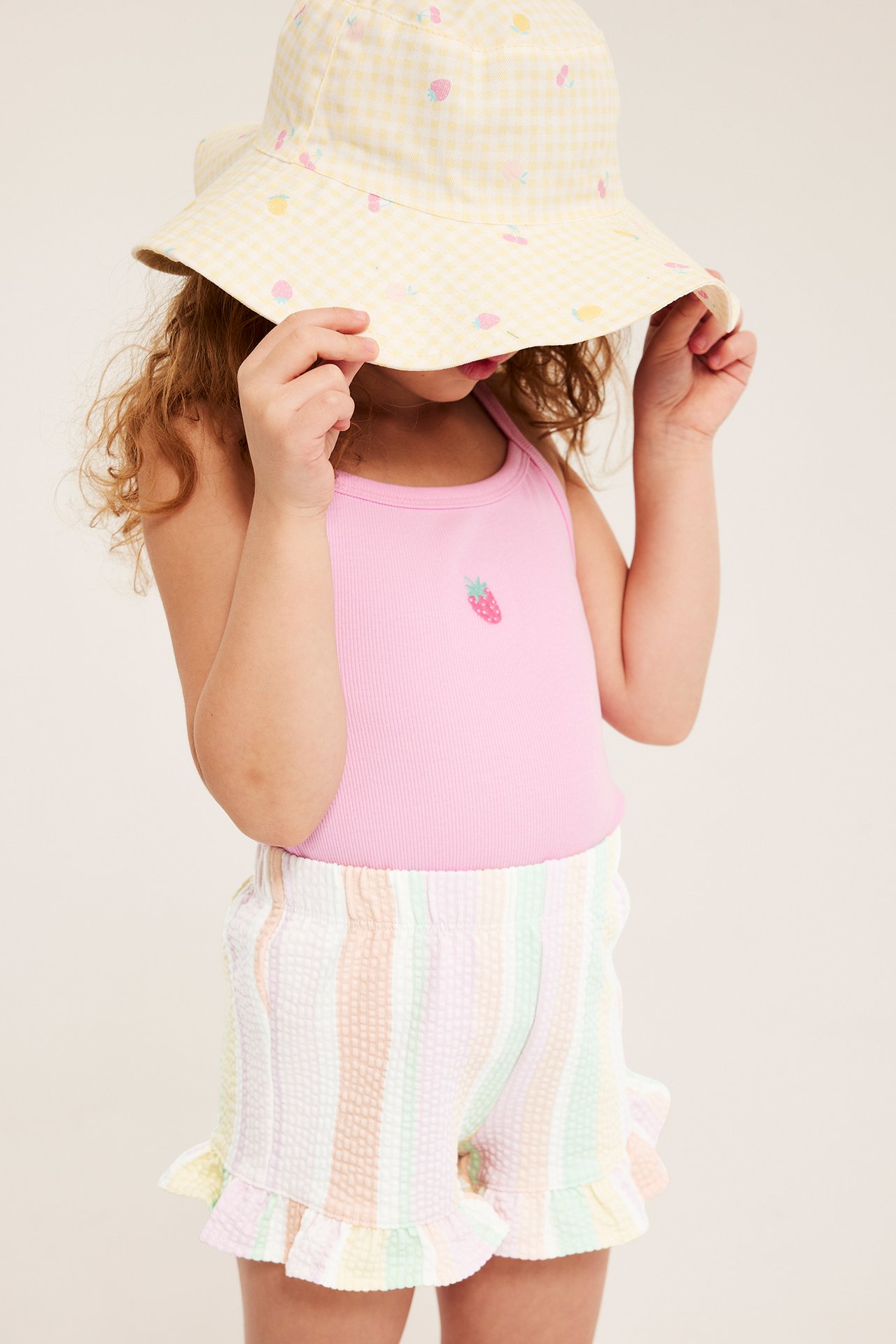 Barn bär flerfärgade randiga bomullsshorts, rosa linne och gul frukthatt.