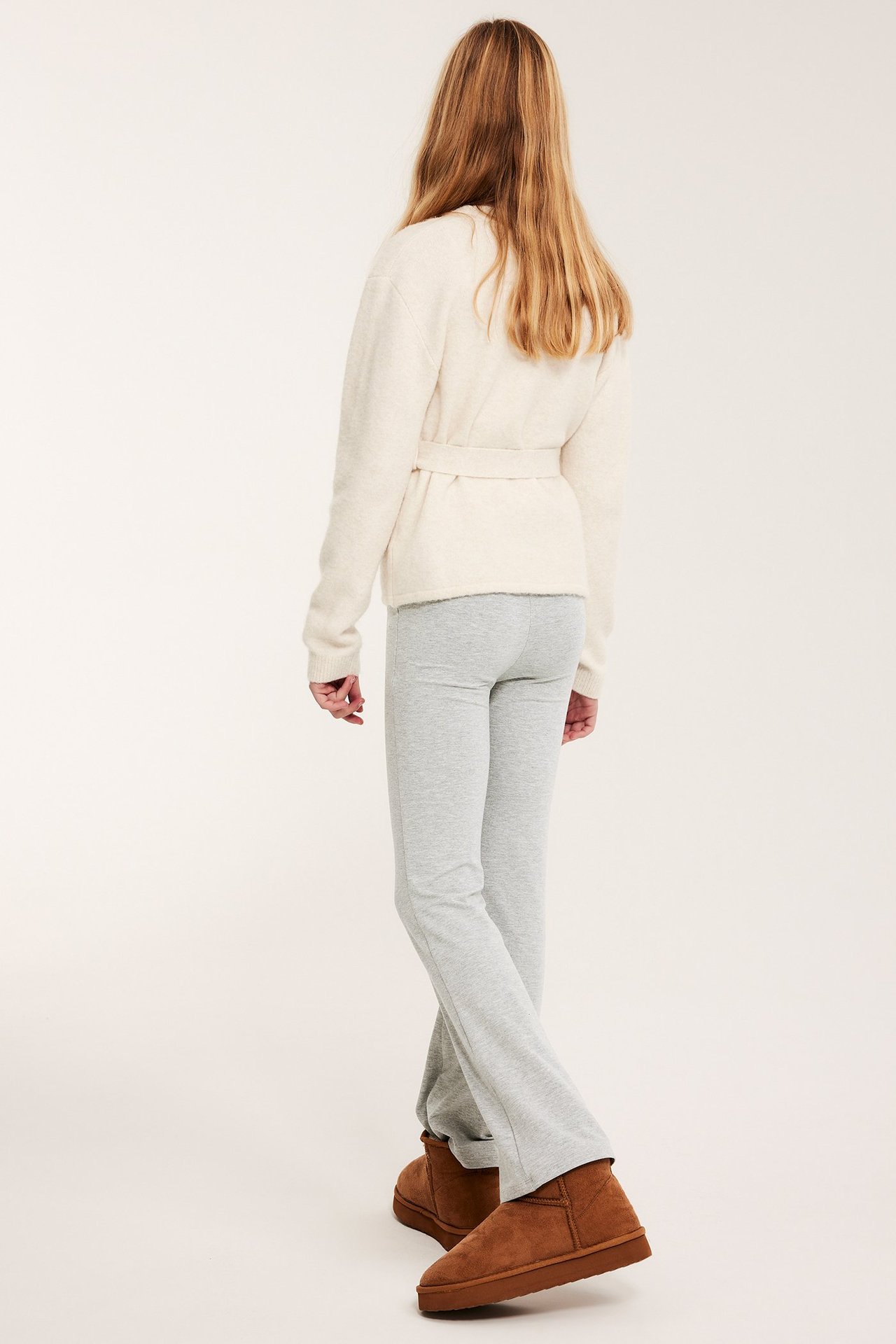 Lapsi takaa: vaaleanharmaat meleeratut levenevät leggingsit, kermanvärinen vyöllinen neuletakki ja ruskeat nilkkurit.