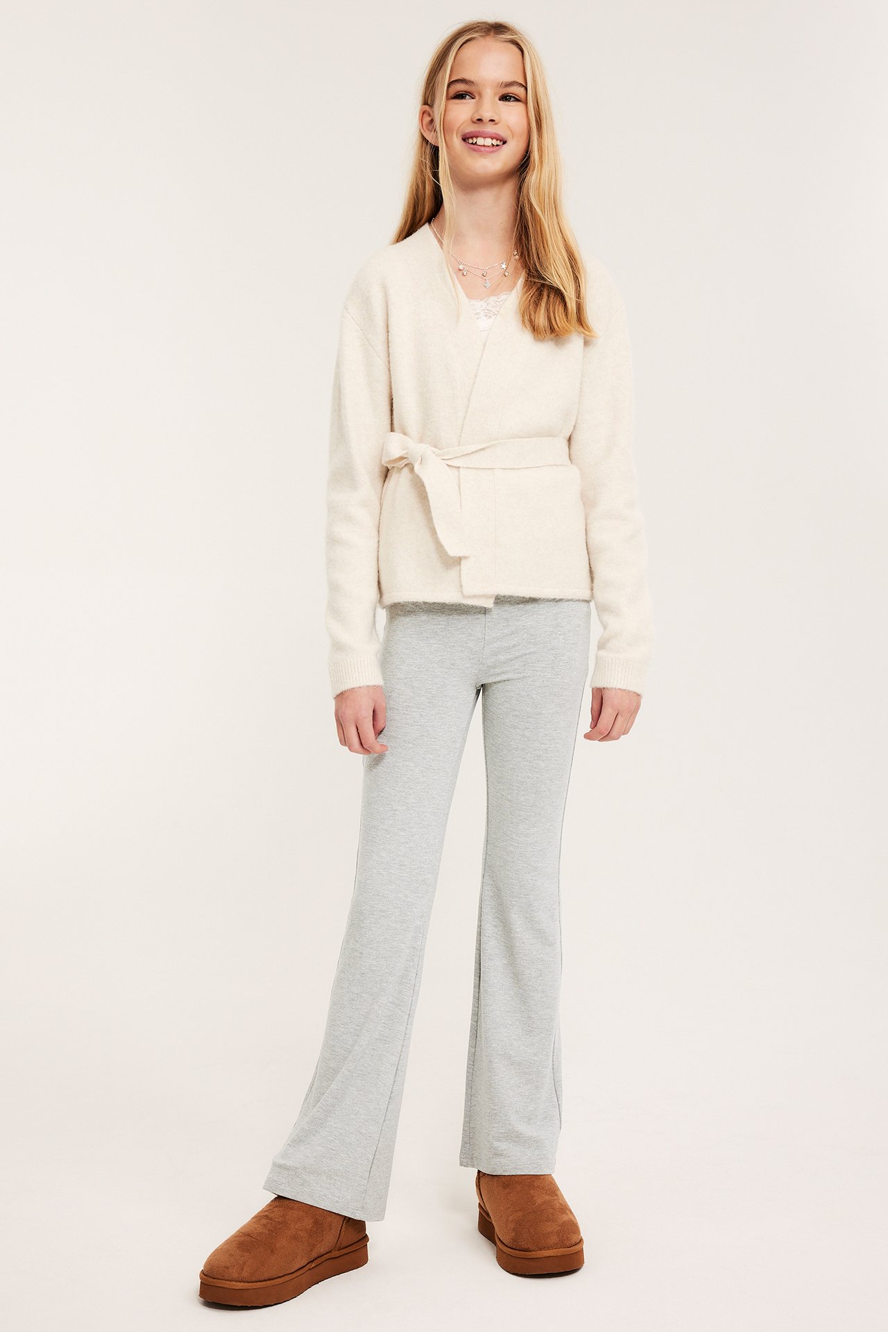 Edestä: Tyttö yllään vaaleanharmaat levenevät joogahousut, luonnonvalkoinen solmittava neuletakki ja ruskeat kengät.